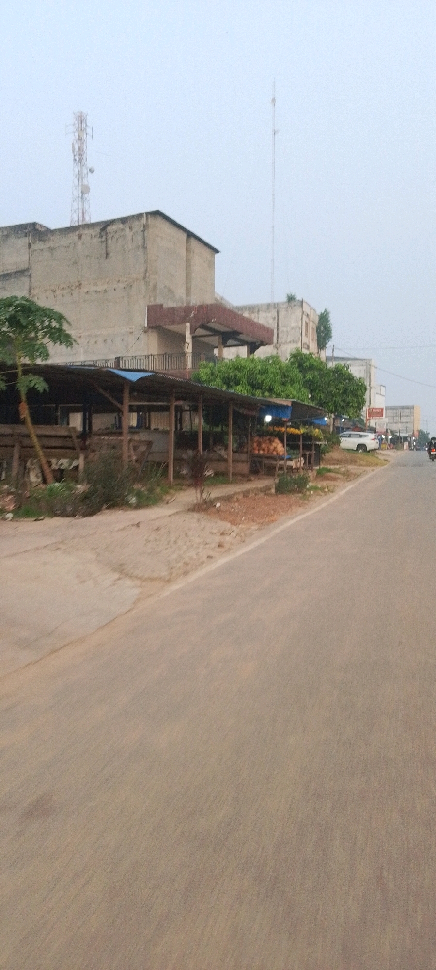 The image shows a roadside scene in Lenggadai Hulu, Indonesia. A grey asphalt road runs diagonally from the bottom right to the upper left of the frame. On the left side of the road, a long, open-air market stall with a corrugated metal roof supported by wooden posts is visible. The stall appears to be selling fruits and vegetables, with piles of coconuts and other produce displayed. Behind the market stall, a large, two-story concrete building with a faded facade dominates the background. A smaller, attached structure with a reddish-brown awning protrudes from this building. Several green trees are planted between the market and the building. Further down the road, other buildings and structures can be seen in the distance, along with a white SUV parked on the right side of the road. A telecommunications tower is visible in the upper left quadrant of the image, rising above the buildings. The sky is overcast with a light grey hue. The overall impression is one of a semi-urban or rural commercial area.