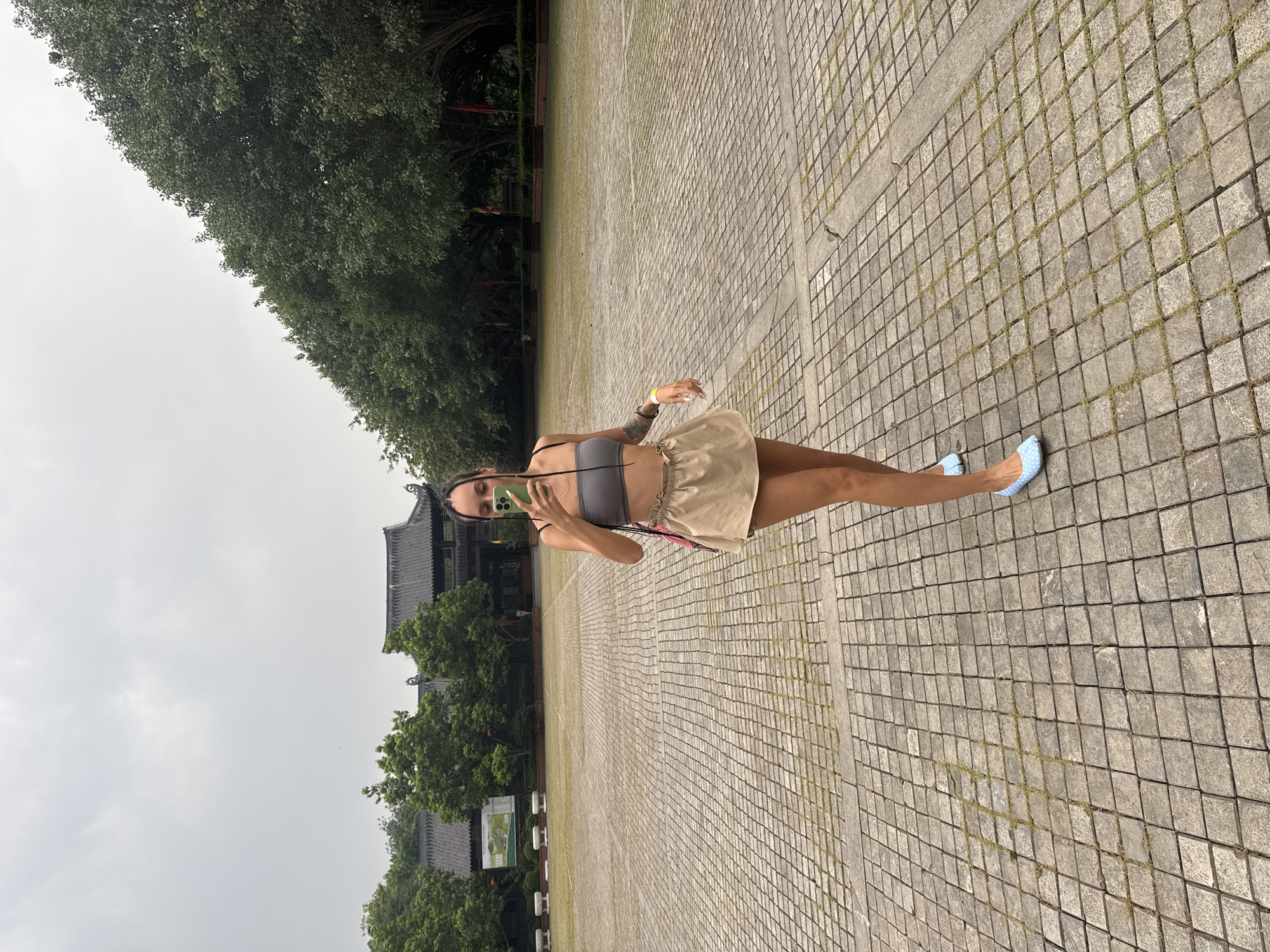 A young woman is taking a selfie with her phone in a large, open courtyard paved with square stone tiles. She is wearing a grey bandeau top, a tan mini-skirt, and light blue polka-dot ballet flats. Her hair is styled in braids, and she has a pink strap over her shoulder. Behind her, there is a traditional Asian-style building with a dark tiled roof and surrounding greenery. The sky is overcast. The courtyard appears to be part of a historical or cultural site. While Tây Hoa Lư Ward is known for its historical significance, this specific location cannot be definitively identified as being within that ward based solely on the visual information provided in the image.