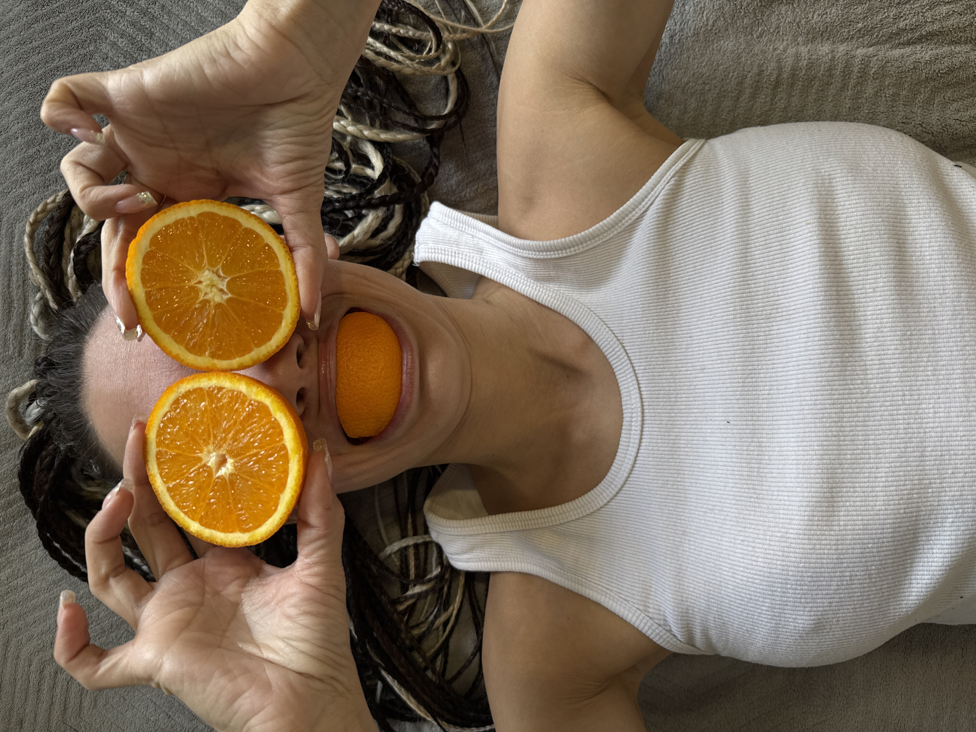 The image presents an overhead view of a person, likely a woman, lying on a grey, textured surface, possibly a bed or blanket, in Rusanovo, Russia. The person is engaged in a creative or humorous pose involving oranges. Two halves of a freshly cut orange are held over their eyes by their hands, resembling oversized sunglasses or eye masks. A third orange half is placed inside their open mouth, humorously substituting for the tongue.
The subject's hands are visible, featuring long fingernails with a light, possibly glittery, polish. Their hair, styled in a striking combination of dark and light braids or dreadlocks, is fanned out around their head. They are wearing a plain white ribbed tank top. The lighting is soft and even, suggesting an indoor setting. The vibrant orange color of the fruit contrasts sharply with the neutral tones of the clothing and background. The overall mood conveyed is playful and artistic. No text is visible in the image.