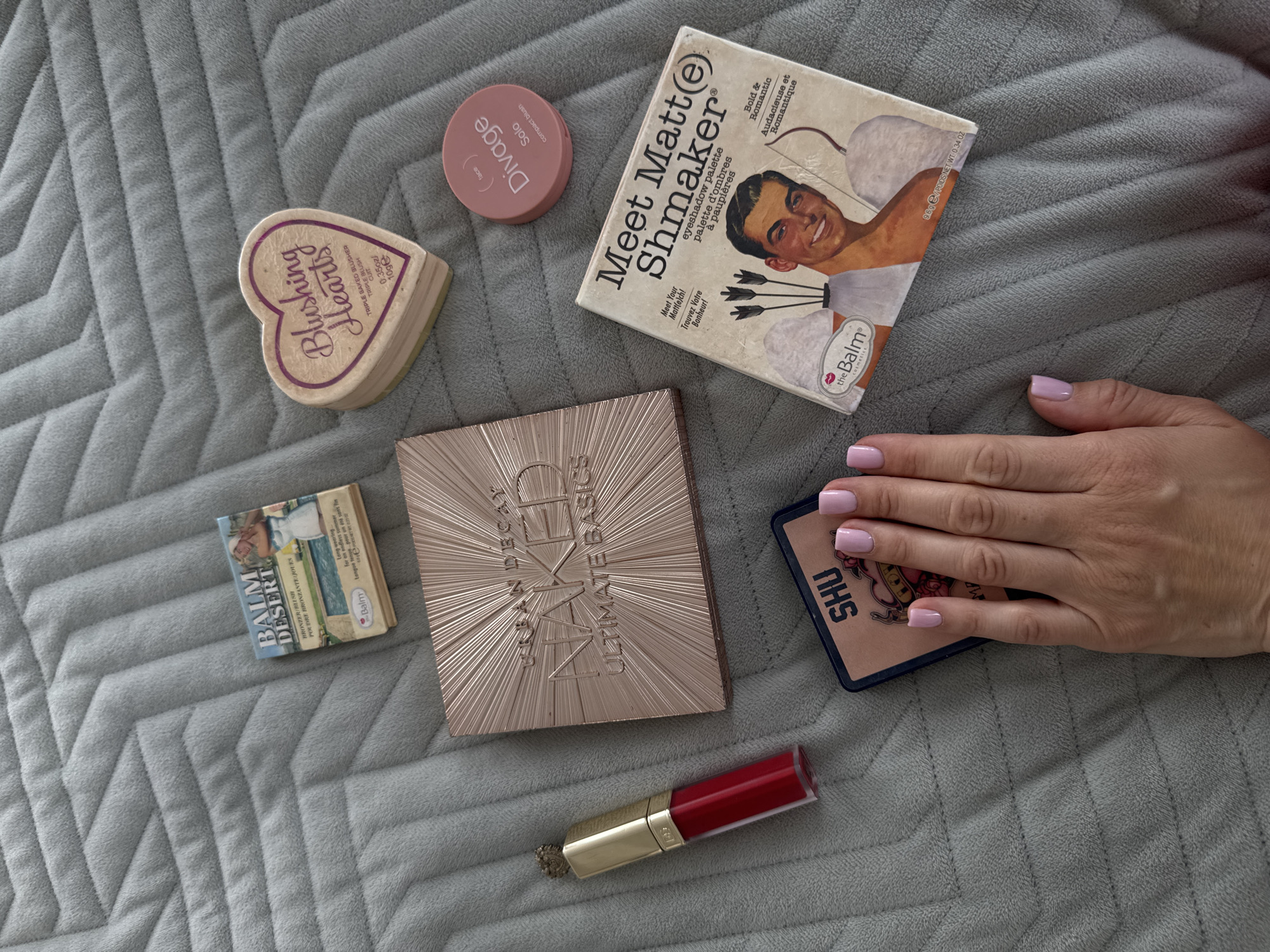 The image shows a collection of makeup products laid out on a gray, quilted surface. The products include a heart-shaped blush compact labeled "Blushing Hearts", a circular blush compact labeled "Divage Solo", an eyeshadow palette titled "Meet Matt(e) Shmaker" by theBalm, a bronzer compact titled "BALM DESERT", an eyeshadow palette labeled "NAKED ULTIMATE BASICS" by Urban Decay, a square compact labeled "SHU", and a red lip gloss tube. A woman's hand with pink nail polish is partially visible on the right side of the image, near the "SHU" compact.
The image does not contain any information or visual cues to determine its location, specifically whether it is in Sochi, Russia.