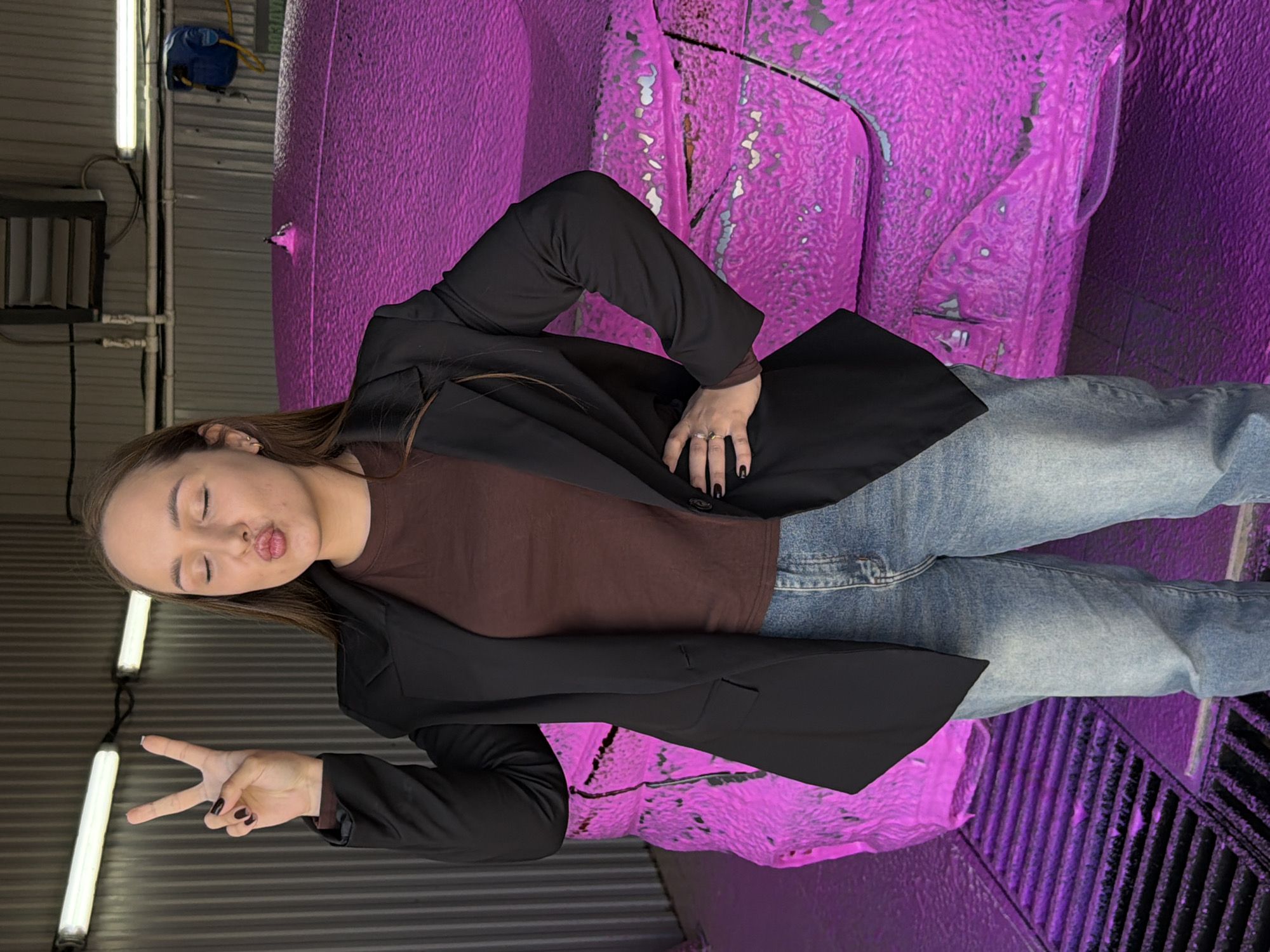 A person stands in what appears to be a car wash. The person is wearing a brown shirt under a black blazer, and blue jeans. They are holding up a peace sign with their left hand and puckering their lips. Behind the person is a partially washed vehicle covered in pink soap. The location appears to be an interior space with fluorescent lighting and ribbed metal walls and flooring. It's hard to place the location specifically within Chaikovsky, Russia, without further visual cues.