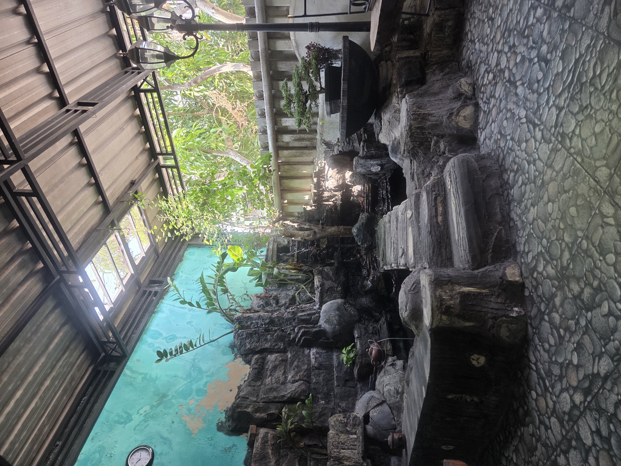 This image depicts an outdoor space, possibly a cafe or a residential backyard, in Sidoarjo, Indonesia. The scene is dominated by a man-made rock garden with a cascading water feature. The rocks are dark gray and textured, arranged to resemble a natural cliff face with several levels and small ledges. Green plants, including what appear to be banana leaves and smaller foliage, are integrated into the rock formation and spill over its edges.
A pathway made of large, flat, gray stones leads across the water feature. The ground is covered with a mosaic of small, rounded stones in varying shades of gray and brown, resembling pebbles. To the left of the rock garden, a light blue wall with patches of peeling paint forms the backdrop. A round clock with a white face and black hands is mounted on this wall.
To the right of the rock garden, a large, dark bowl contains a bonsai tree with lush green foliage. Beside the bonsai, a smaller plant with dark red leaves is also visible. Behind the rock garden and bowl is a light-colored concrete wall with a decorative pattern of circular indentations. Beyond this wall, trees with abundant green leaves are visible, suggesting a lush, tropical environment.
A black lamppost with three glass lantern fixtures stands to the right of the scene, casting light downwards. Part of a wooden bench or table is visible to the far right. The space is covered by a sloped roof constructed of corrugated metal sheets supported by a dark metal frame. Several sections of the roof are open, allowing sunlight to stream through, illuminating the scene. The overall impression is one of a tranquil, garden-like setting designed for relaxation or social gathering.