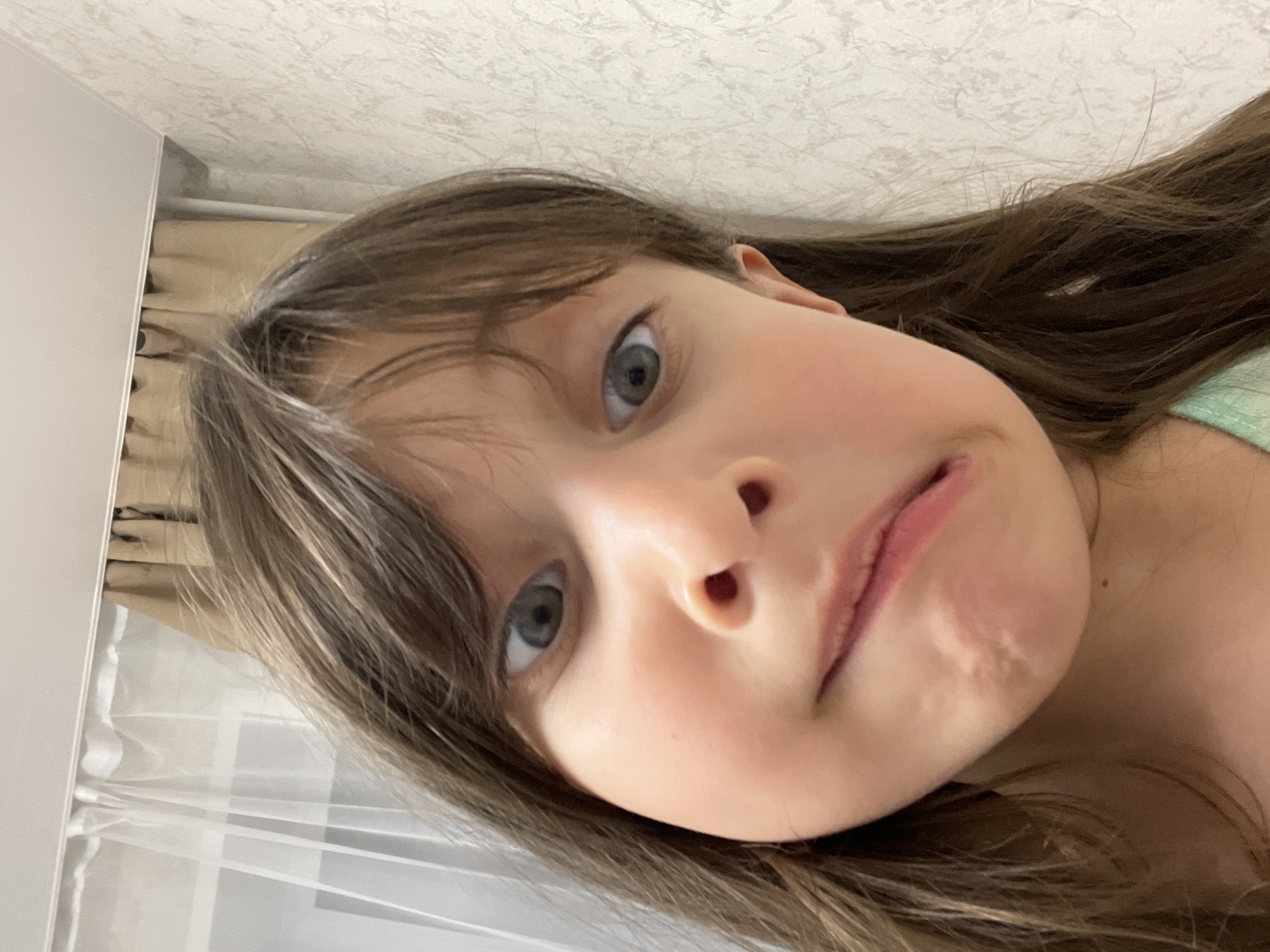 A close-up, slightly angled portrait of a young girl with light brown hair and blue eyes. She has fair skin and a slightly puckered expression. The background includes a white wall with a textured pattern and a glimpse of a window with white curtains. There is no information in the picture that allows identifying the location in the city of Izhevsk, Russia.