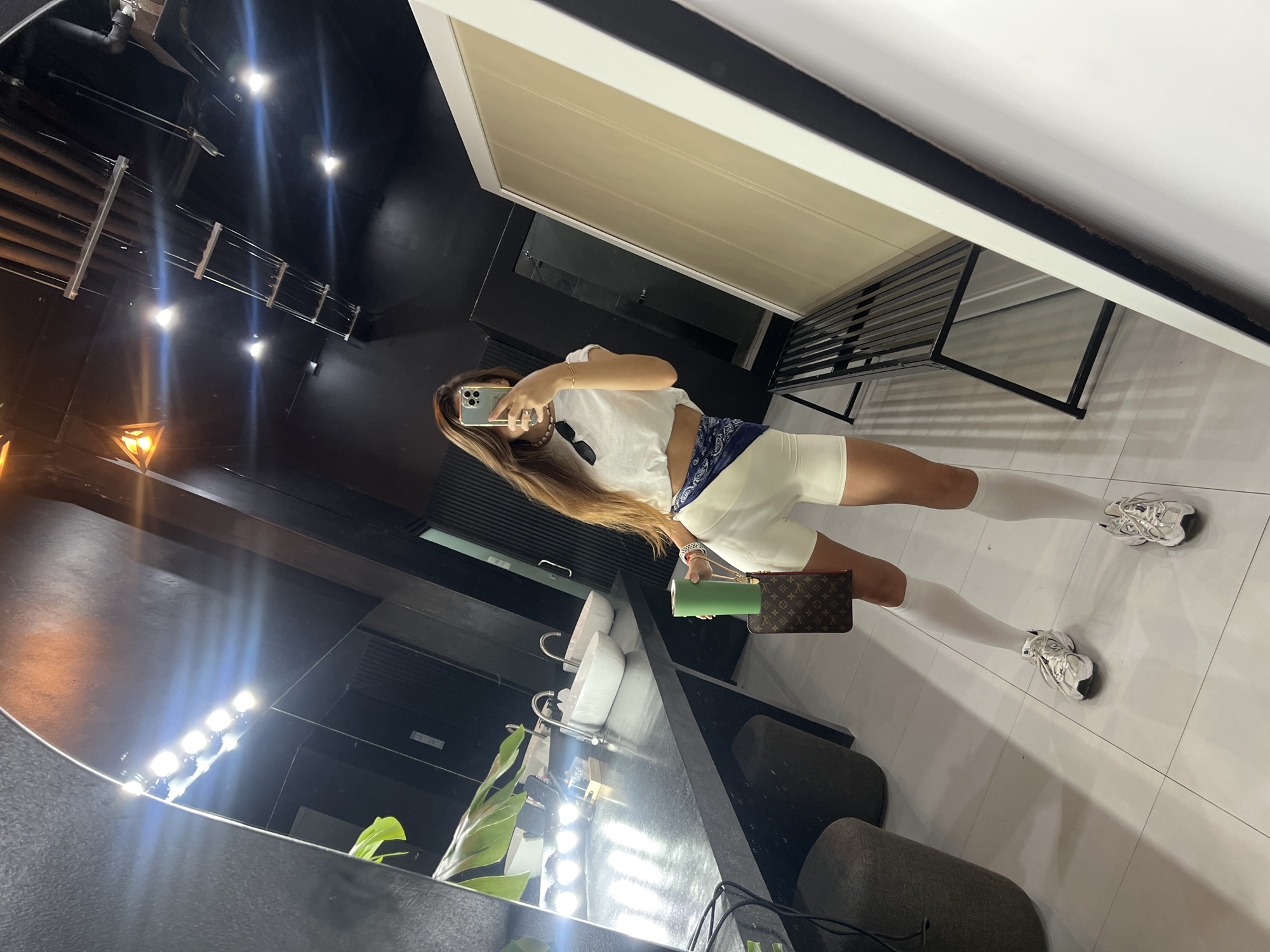 A person is taking a selfie in a modern, minimalist bathroom with a large mirror. The person is wearing a white crop top, white biker shorts, white knee-high socks, and white athletic shoes. They are holding a green water bottle and a small brown designer handbag in their left hand. The bathroom features a black countertop with two white sinks, a large mirror with bright lights above it, and black walls and ceiling. There is a plant on the left side of the counter and a black metal bench on the right. The floor is tiled with large white square tiles. The overall aesthetic is clean, sleek, and somewhat industrial.