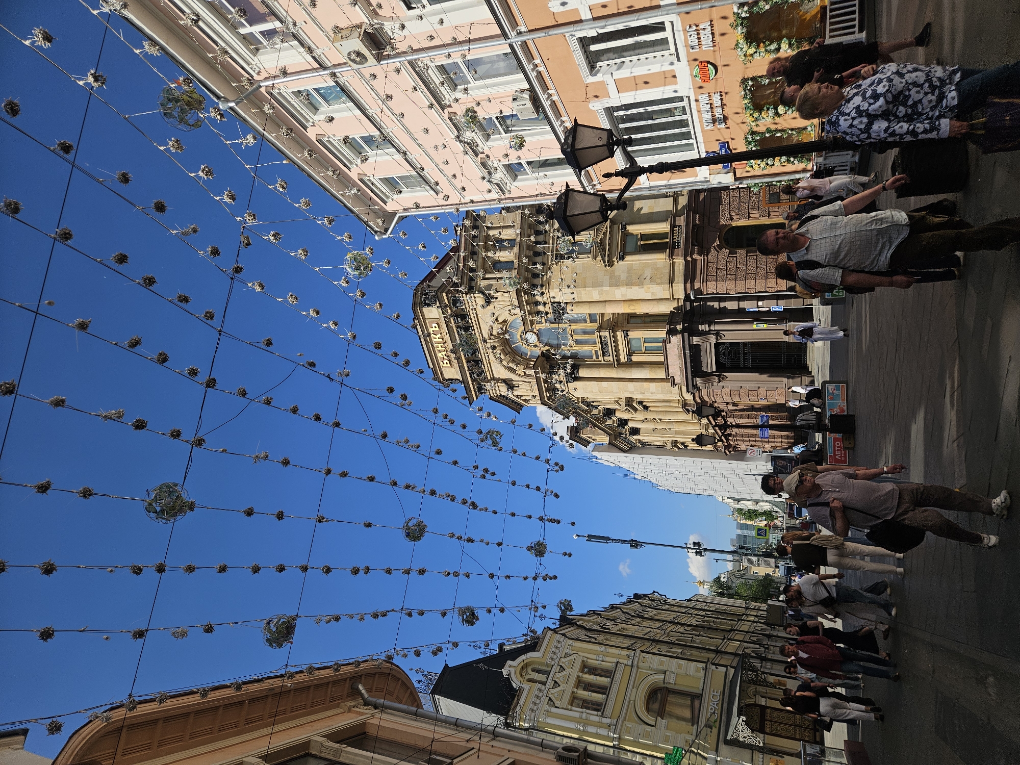 This vibrant daytime street scene captures a bustling urban environment under a clear blue sky. The perspective looks upwards along a street lined with ornate, multi-story buildings. Numerous decorative spherical elements, some simple and spiky, others more elaborate with internal structures, are suspended across the street on a grid of wires, creating an overhead canopy.
The buildings on both sides of the street exhibit classic architectural styles with intricate facades, columns, arched windows, and balconies, rendered in shades of light pink, beige, and earthy tones. Ornate black streetlights are also visible, contributing to the historical feel of the area.
Several pedestrians are walking along the wide sidewalk on the right side of the street. In the mid-ground, a man in a light plaid shirt, a cap, and a shoulder bag is prominent, walking away from the camera. Other individuals, including a person in a floral top and others in casual attire, are seen strolling further down the street, engaging in everyday activities.
Visible text on storefronts and signs is in Cyrillic script. On a beige building, the word "БАНКЪ" (BANK) is clearly readable above an entrance. Further down the street, partial words like "ЛЕТО" (LETO - Summer), "ГИПС С..." and "ТАБА" can be seen on various signs, indicating commercial establishments. The overall atmosphere suggests a lively pedestrian street, likely in an Eastern European city.