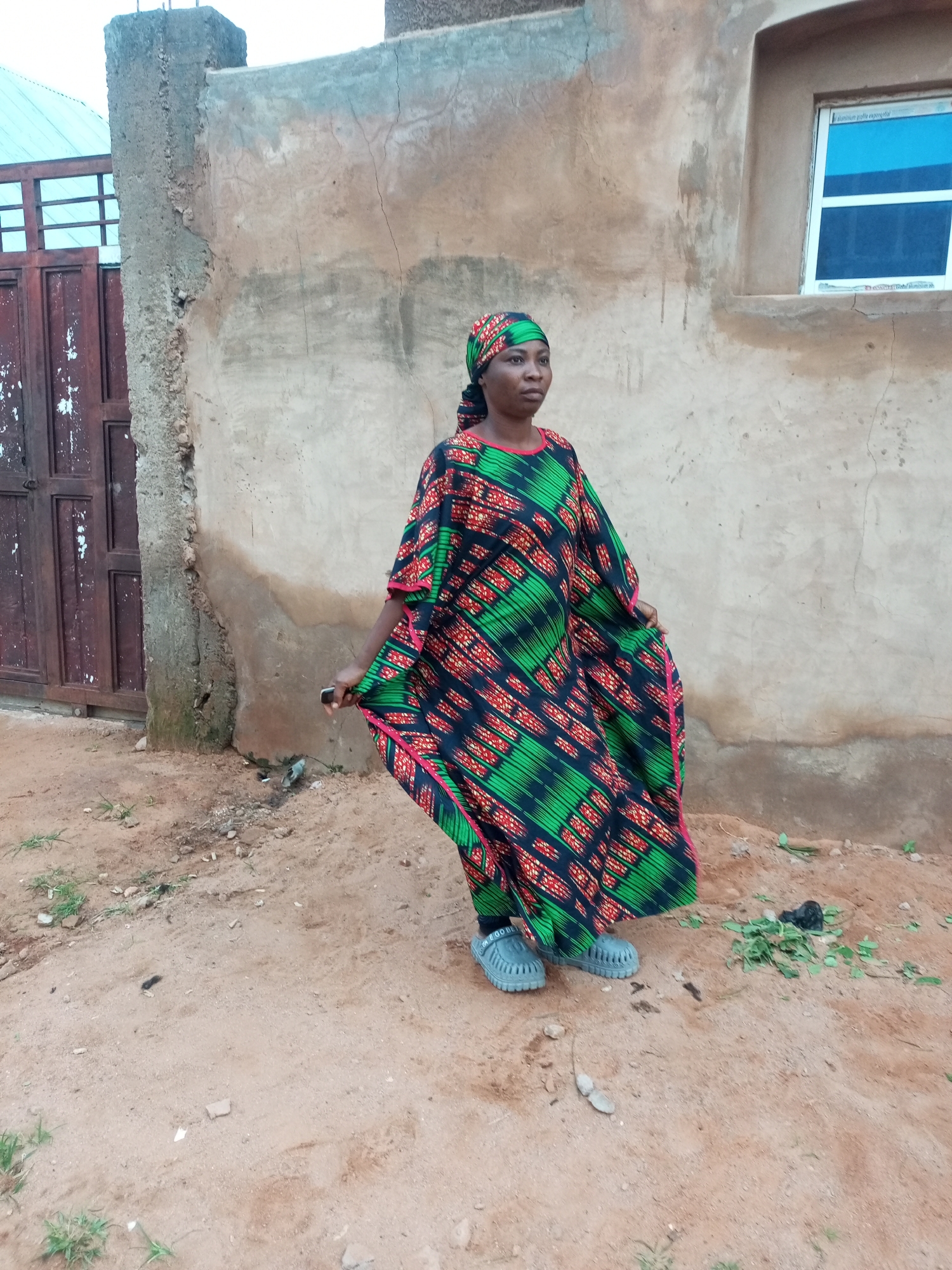 A full-body, eye-level, outdoor medium shot shows a woman standing in front of a light brown, stucco-walled building in Tempure, Nigeria. The woman, who is facing to her right, is wearing a dark green, red, and black patterned kaftan with a matching headscarf. She is also wearing gray slip-on shoes. Her right hand is holding the fabric of the kaftan. The building has a single, blue-paned window with a white frame on the right side of the image. To the left of the woman, there is a dark brown, metal double door with a lattice pattern. The ground in front of the building is covered in dirt with some patches of dry grass and scattered debris. The lighting suggests it is daytime, and the sky is overcast.