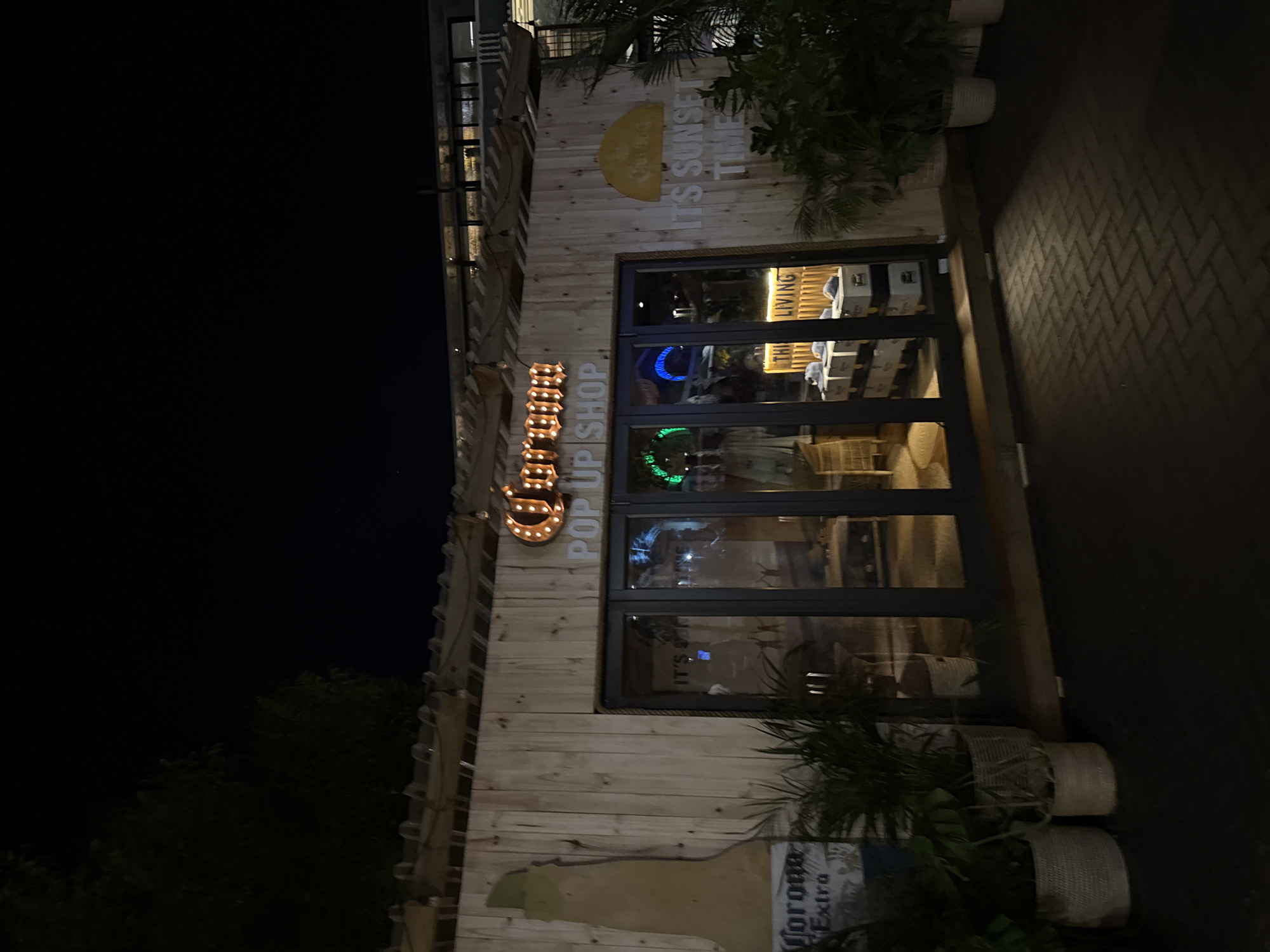 A Corona Pop Up Shop is depicted at night in Cape Town, South Africa. The shop's exterior features a light-toned vertical wooden plank facade under a flat roof with exposed wooden beams, from which string lights are suspended. Above the central dark-framed glass bi-fold doors, an illuminated "Corona" sign in marquee-style letters is prominently displayed, positioned directly above the text "POP UP SHOP." To the left of the entrance, a large cutout of a Corona Extra beer bottle is affixed to the wooden wall. To the right, a yellow semicircular logo is present, accompanied by the text "IT'S SUNSET TIME."
Through the four glass door panels, the brightly lit interior is visible, showcasing light-colored baseball caps arranged on white crates or stands. A partially visible sign inside reads "THI LIVING." Interior lighting includes prominent green and blue illumination. The shop's front is adorned with multiple potted plants contained within light-colored woven baskets or pots. The ground in the foreground consists of dark, herringbone-patterned brick paving. Above the shop, the sky is dark, with the silhouette of tree foliage visible in the upper left quadrant of the image. No individuals are present within the scene.