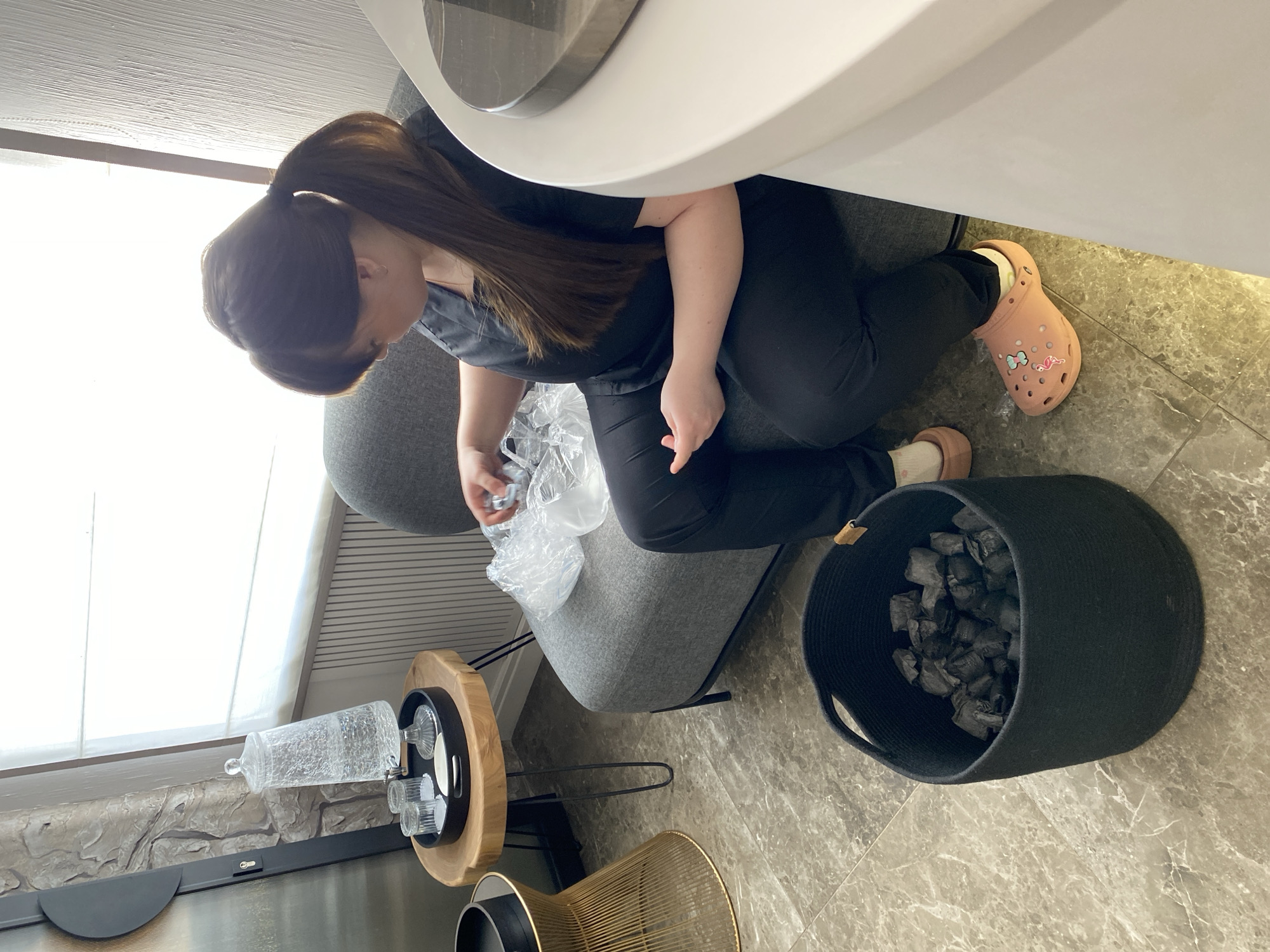 A woman with brown hair in a ponytail, wearing black clothing and salmon-colored Crocs, kneels next to a gray chair and is unwrapping something. Next to the chair is a small, round wooden table holding clear glasses, a tray, and a glass container, as well as a gold wire stool. To the right of the chair is a black basket filled with black trash bags. The floor is gray tile. The location cannot be determined from the image but has been specified as Votkinsk, Russia.