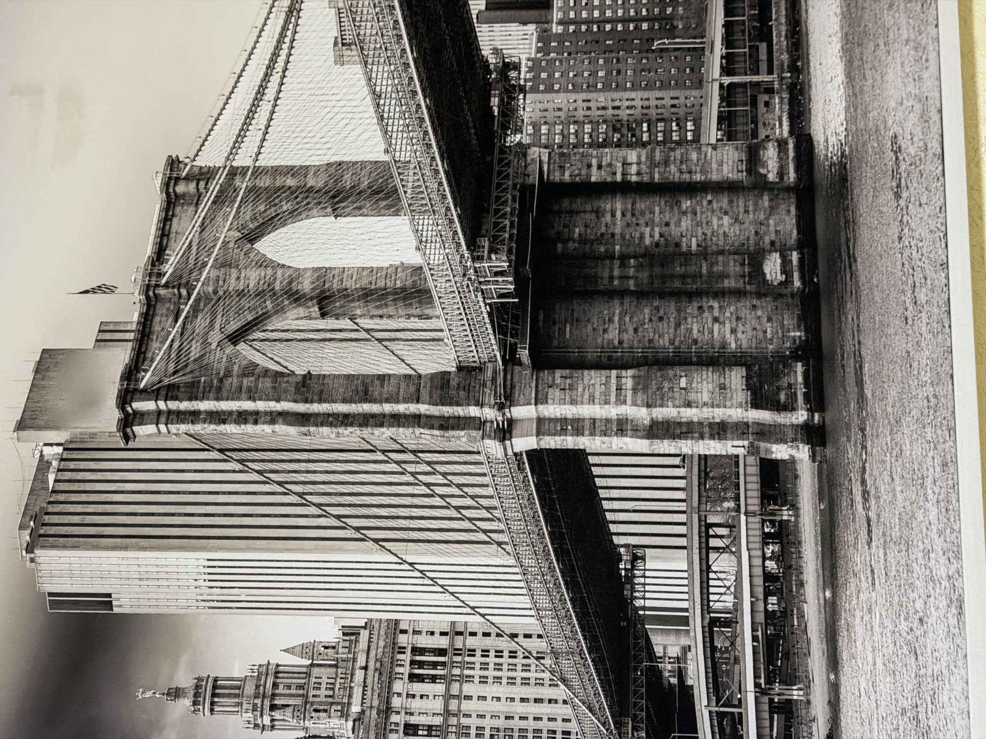 The image is a black and white photograph of the Brooklyn Bridge in New York City. In the foreground, the East River is visible with its textured surface reflecting light. The iconic stone towers of the Brooklyn Bridge dominate the midground, with their massive arches and intricate suspension cables stretching across the frame. The walkway of the bridge is clearly depicted, showing its metal railings and structure. Behind the bridge, a cluster of tall buildings rises into the sky, including a distinctive art deco building on the left with a spire and a statue. The background features a hazy sky and more urban development, with apartment buildings lining the waterfront. The photograph is a classic cityscape, emphasizing the architectural grandeur of the bridge and its urban surroundings.