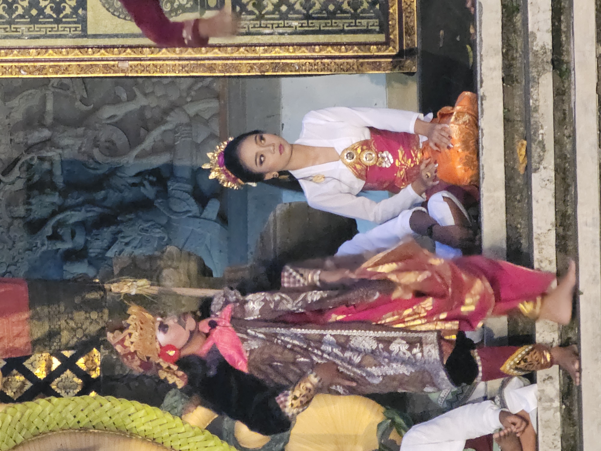 A Balinese dance performance is taking place on a stage with stone steps. Two dancers are prominently featured. On the left, a dancer in a black top, elaborate headdress, and ornate skirt and leggings, likely a male dancer due to the facial makeup. On the right, a female dancer sits with her hands on a piece of orange fabric, wearing a white top, a pink and gold patterned skirt, and a golden headdress. The background features a carved stone structure and a golden ornate frame. The scene appears to be outdoors, possibly in a temple complex or cultural venue within Denpasar, Indonesia, given the traditional attire and performance style.