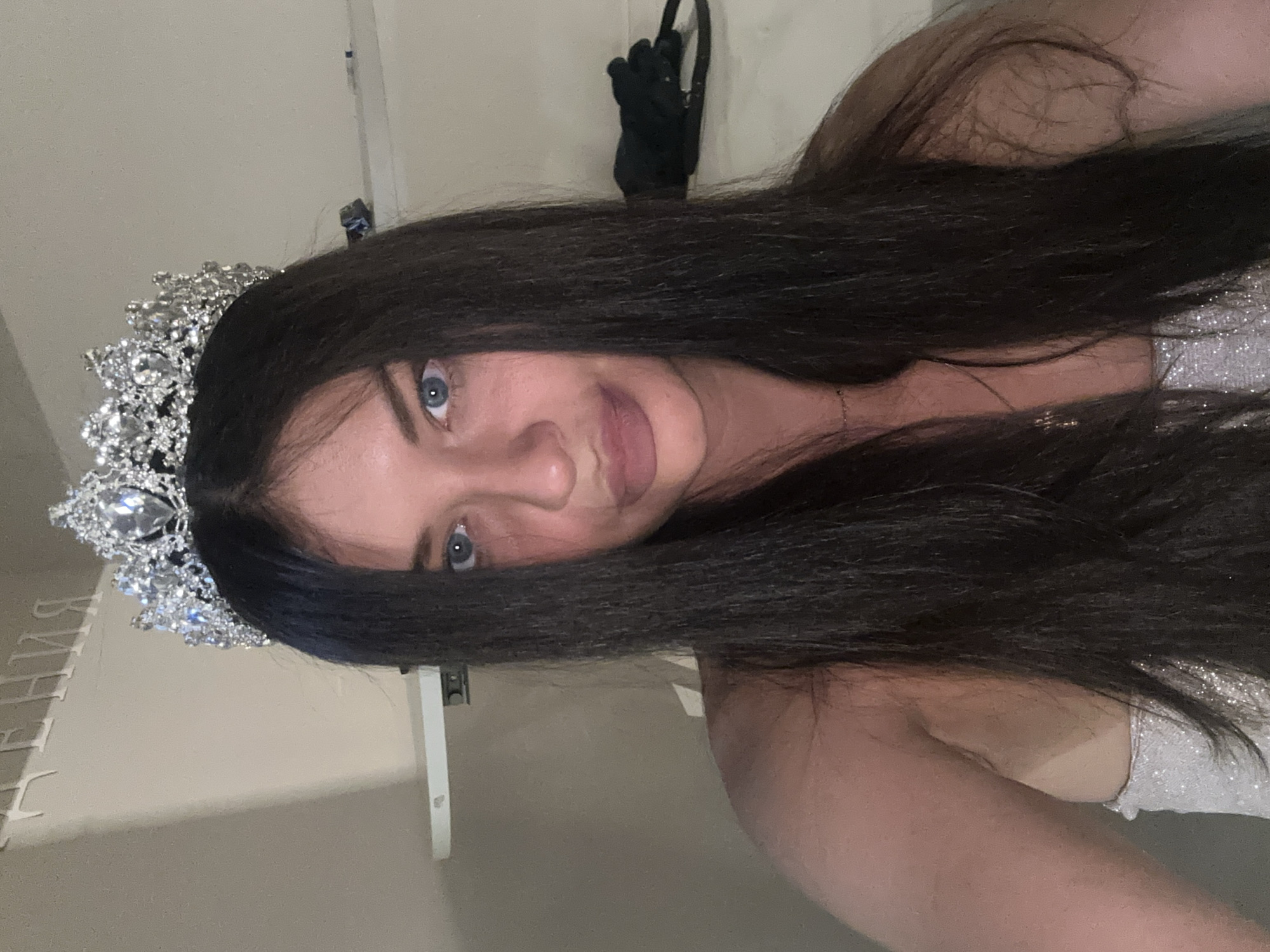 The image, rotated 90 degrees counter-clockwise, displays a young woman with long, dark hair, looking directly at the camera. She wears a large, ornate silver-colored tiara or crown, lavishly adorned with numerous sparkling clear jewels. Her eyes are blue, and she exhibits a slight, closed-mouth smile, conveying a serene and pleasant expression. Her dark hair flows over her shoulders, and a glimpse of a light-colored, possibly sparkly garment is visible on her upper body.
The setting is indoors, with a plain, light-colored wall serving as the background. In the upper right, a black object, possibly a hanging glove or accessory on a hook, is visible. White vertical elements, likely part of shelves or furniture, are present in the background on both sides.
The woman appears to be posing for a photograph, perhaps a selfie, suggesting a celebratory or formal event, such as a pageant or special occasion, emphasized by the ceremonial crown. In the lower-left background, partial Cyrillic text is visible, specifically "РЕНИЯ" with the preceding letters obscured, possibly part of a larger word or title. This scene takes place in Moscow, Russia.