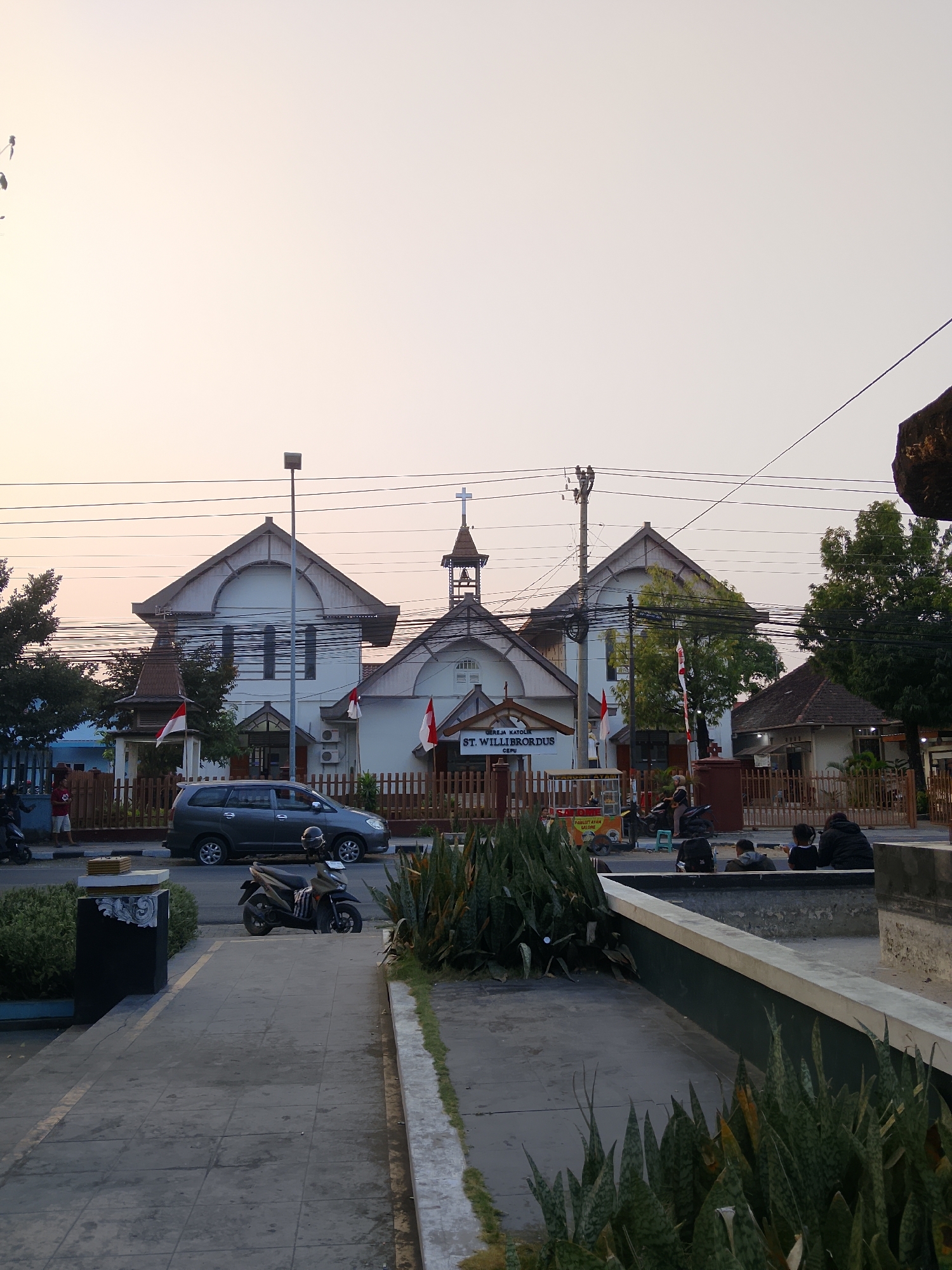 The image depicts the St. Willibrordus Catholic Church in Balun, Indonesia, under a pale sky with the sun low on the horizon, casting a warm, soft light. The church features a central bell tower topped with a cross and two adjacent buildings with distinctive gabled roofs. Indonesian flags are displayed prominently on flagpoles in front of the church, suggesting a national holiday or commemoration. A paved walkway leads to the church, with a dark-colored minivan parked on the adjacent street. A scooter is also parked on the sidewalk, and several individuals are visible in the background, including a man standing near a fence and a group of people sitting by a fountain. Lush green plants, including snake plants, are in the foreground, adding a touch of nature to the urban landscape. The overall atmosphere is serene and peaceful, capturing a quiet moment in the life of the community.