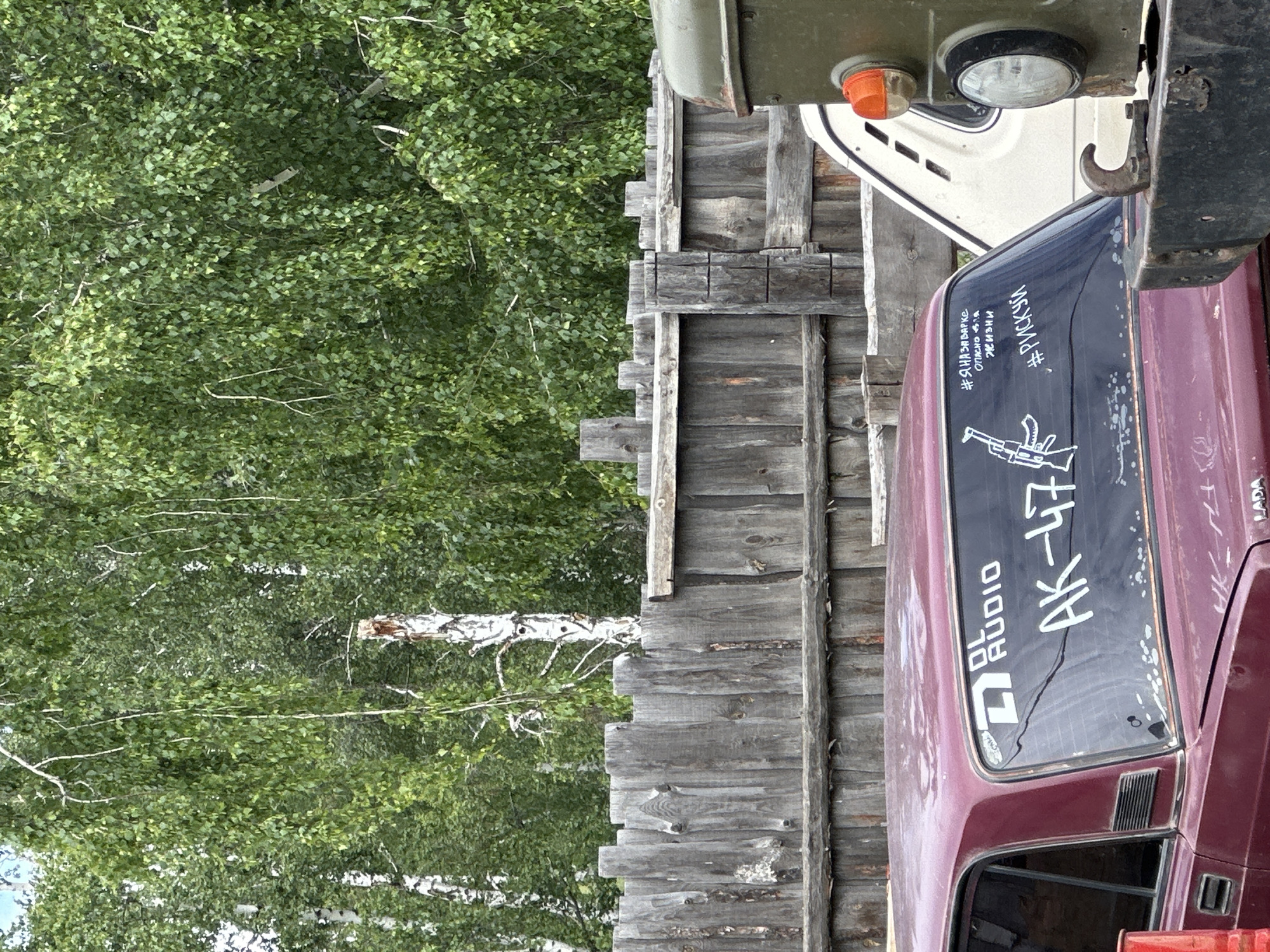 An outdoor scene in Unknown, Unknown, captures a maroon Lada sedan and a partial view of an olive green utility truck behind a weathered wooden fence. The Lada is positioned with its rear facing the viewer, featuring white stickers on its back window. These stickers include a "DL AUDIO" logo, the text "AK-47" accompanied by a white outline drawing of an AK-47 assault rifle, and two Cyrillic hashtags: "#Я НА ЗВУКЕ ОПАСНО ДЛЯ ЖИЗНИ" (translating to "#I AM ON THE SOUND SYSTEM DANGEROUS FOR LIFE") and "#РИСКУЙ" (translating to "#RISK IT"). The Lada shows signs of wear, including faded paint and some imperfections on the rear window and bodywork. To the right, the front section of a heavy-duty, olive green vehicle is visible, including a prominent metal bumper with a tow hook, a circular headlight, and an amber turn signal. Behind the vehicles, a rustic wooden fence, composed of vertical, unpainted planks, spans the midground. Beyond the fence, a dense collection of birch trees with vibrant green leaves forms the background, suggesting a forest edge or a wooded area. The sky above is bright but partially obscured by the tree canopy. No individuals are present in the image.
