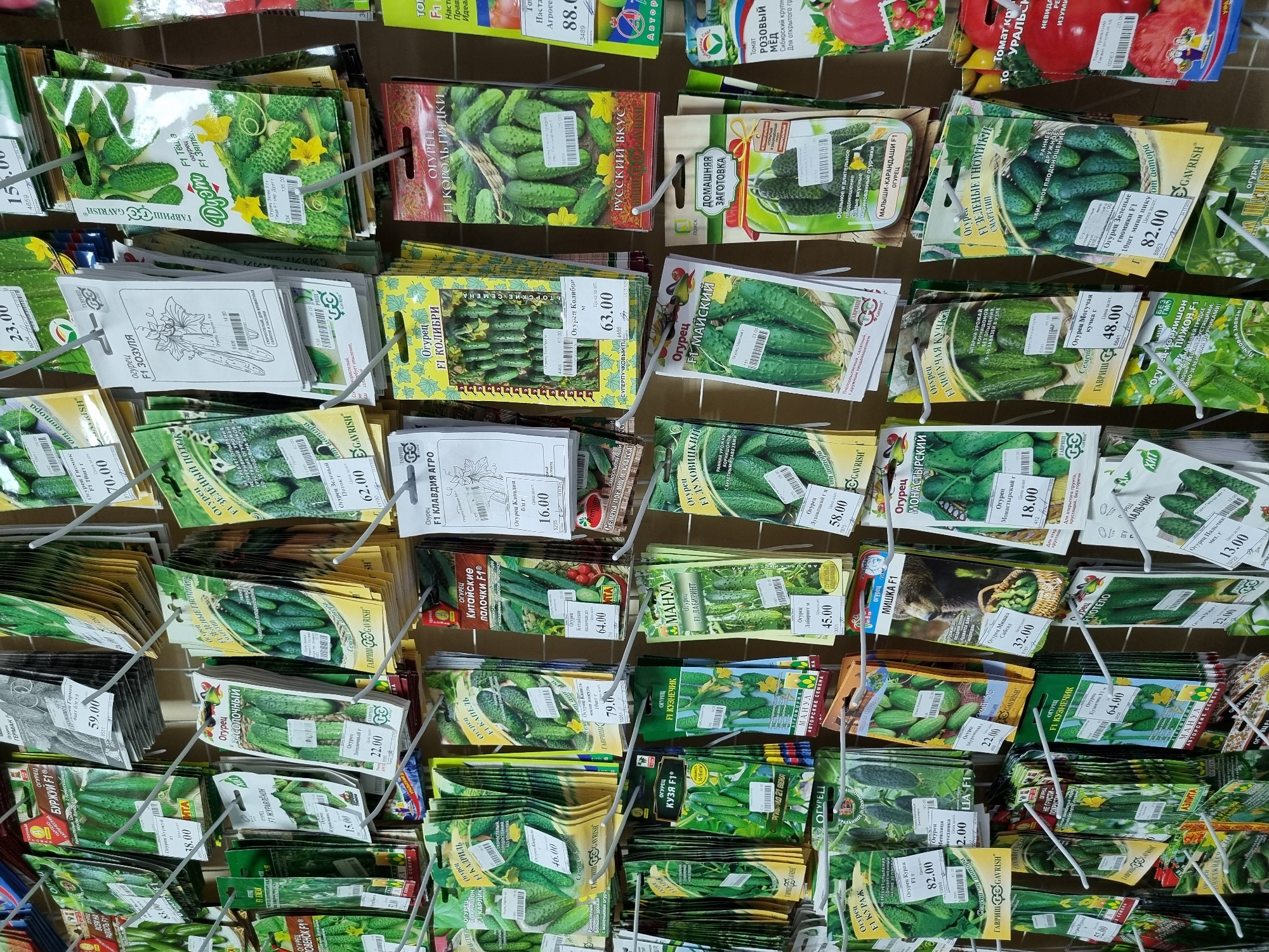 The image shows a display rack filled with various packets of cucumber seeds for sale, likely inside a gardening store or supermarket in Chaikovsky, Russia. Most of the seed packets feature images of different cucumber varieties, including "Burchuy F1," "Geran," "Kuznechik F1," "Zozula," "Maysky," "Kurazh," and many others. Each packet has a price tag affixed, with the prices ranging from 13.00 to 88.00. The packets are hung on metal wire hooks that form the rack, creating a dense and colorful display. Some packets also show tomato seed options.