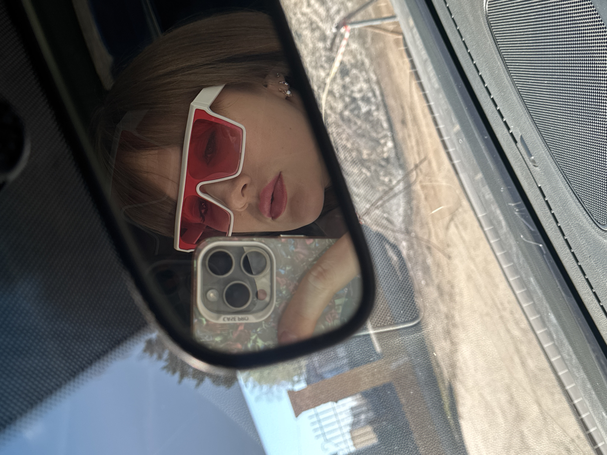A reflection in a car's side mirror shows a person with light brown hair and red sunglasses taking a selfie with a smartphone. The phone has a floral-patterned case. Outside the car, a street with buildings is visible. The scene suggests a location in the city of Chaikovsky, Russia. The surrounding environment includes parts of the car interior such as the window, speaker, and dashboard.
