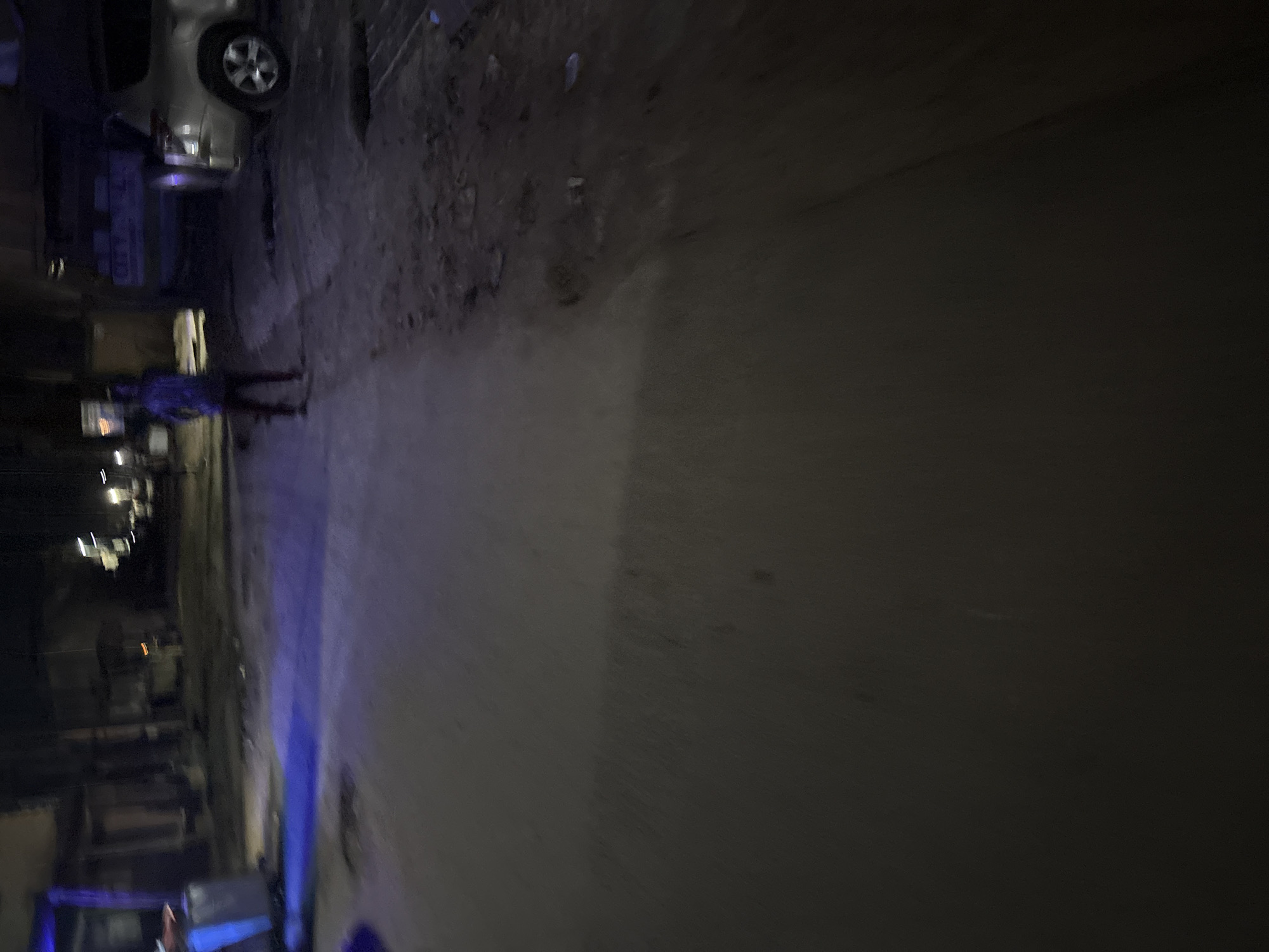 A person stands on a dirt road at night, illuminated by a blue light source. The person is wearing a dark shirt and pants and is facing away from the camera. A light-colored SUV is parked on the right side of the road. The road is unpaved and appears to be in disrepair, with potholes and debris visible. Buildings line the left side of the road, with some appearing to be shops or homes. The overall scene suggests a nighttime street in a developing urban area, possibly within the city of Onitsha, Nigeria, given the commonality of such environments in that region. The lighting indicates that this is likely a low-light or nighttime street scene, with artificial illumination casting shadows and highlighting textures.