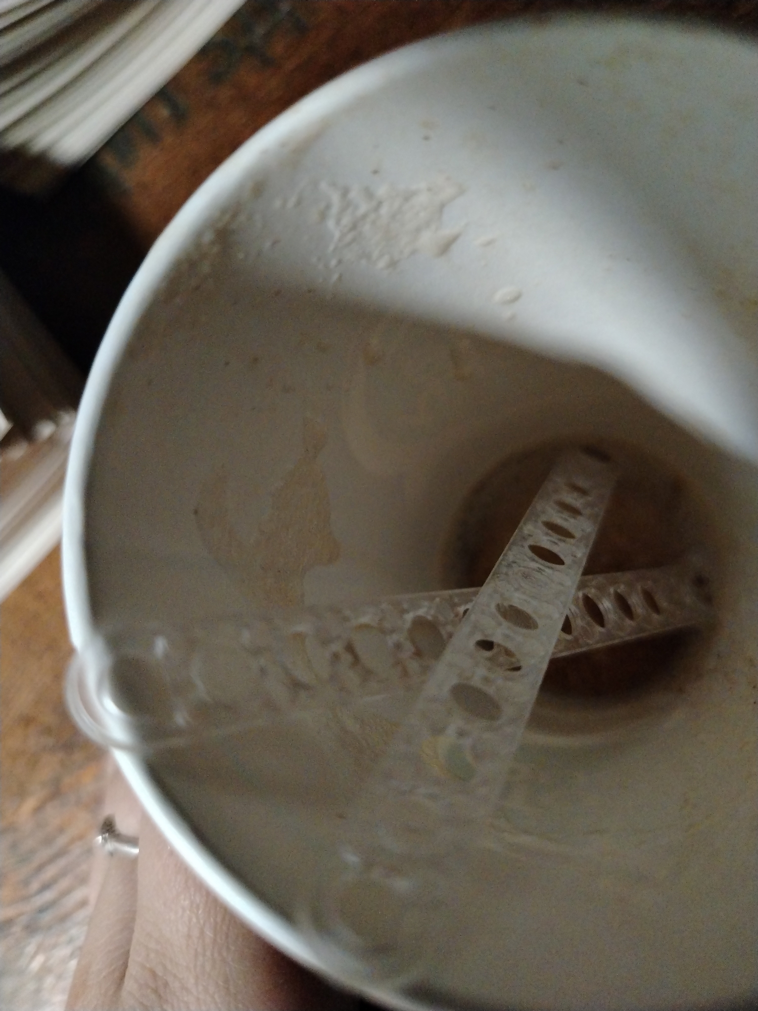The image offers a top-down perspective into a white cup or mug, likely a shaker bottle, held by a hand in the lower left. Inside the cup, resting at the bottom, is a clear or translucent plastic blender ball, formed by two intersecting, perforated strips. The interior surfaces of the cup are smudged with a light brown or beige powdery residue, indicating a recently mixed or consumed beverage, such as a protein shake or powdered drink.
In the blurred background, a stack of papers or books is visible on the left, next to a wooden surface that stretches across the upper and right parts of the frame. The setting appears to be indoors. There is no visible text or identifiable branding within the image. The scene captures the aftermath of preparing a mixed drink.