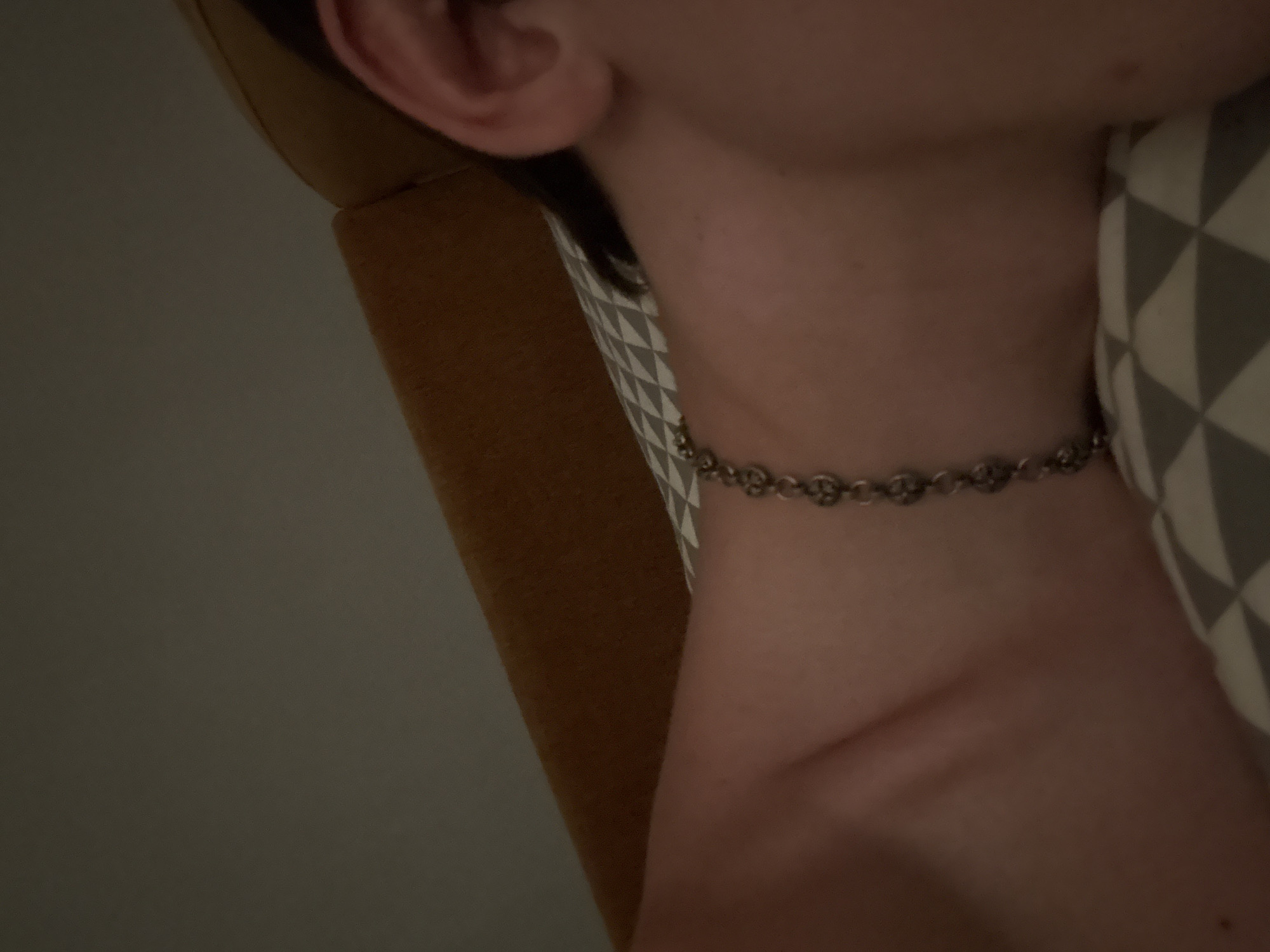 The image shows a close-up of a person's neck and ear, with a silver bracelet visible on the neck. The person appears to be lying down, possibly on a pillow with a geometric pattern. A brown headboard or similar furniture is visible in the background, along with a plain, dimly lit wall. The scene is indoors, and the lighting suggests it might be evening or nighttime. There is no information in the image to indicate the location within the city of Izobilnyy, Russia.