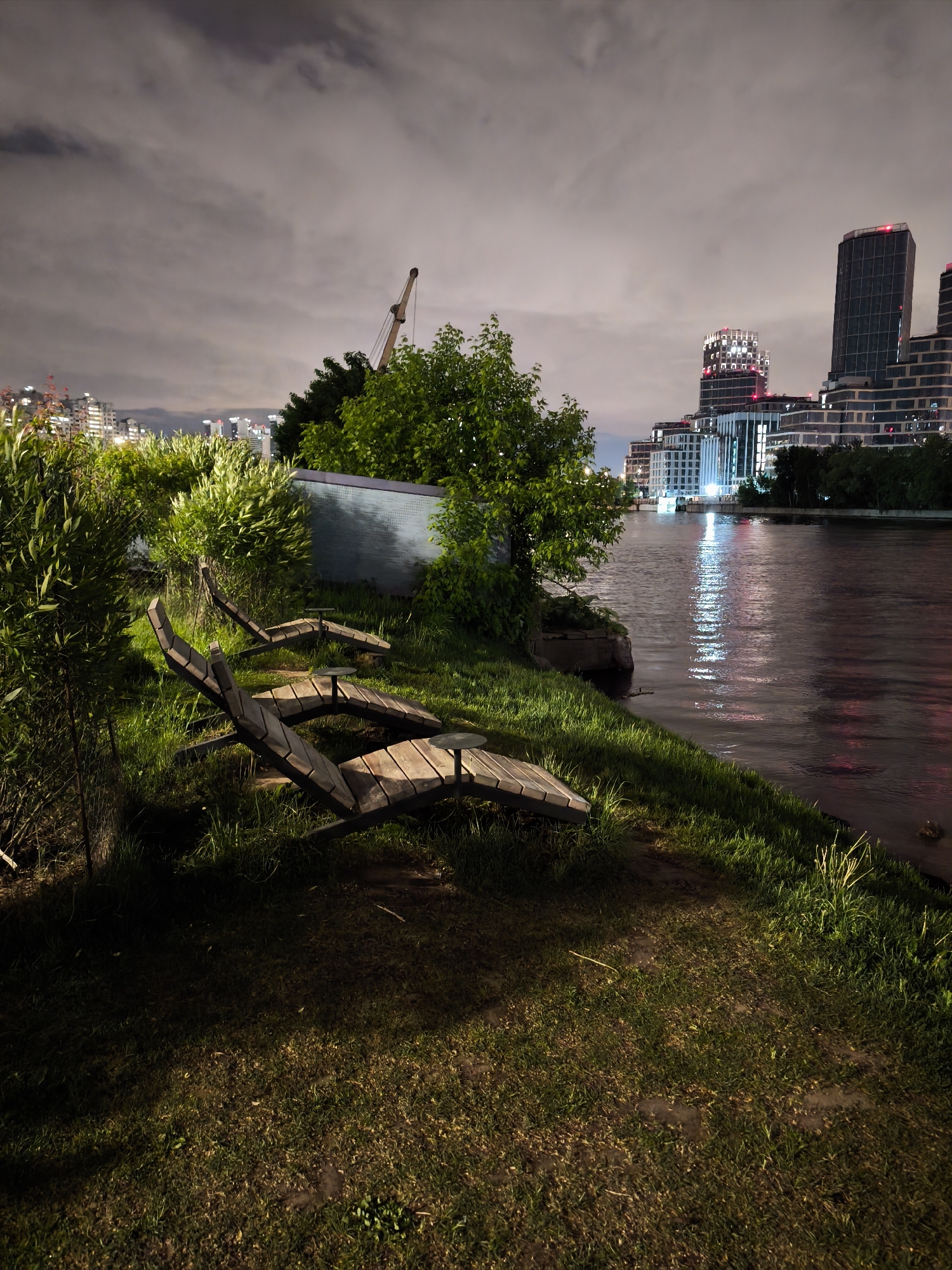The image shows a waterside park area in Moscow, Russia, at night. Two empty wooden lounge chairs are positioned on a grassy embankment. A low concrete structure with writing is partially obscured by bushes. Water reflects city lights from tall buildings on the opposite bank. The sky is overcast. A construction crane is visible among the trees. The light is dim, with highlights from the city illumination. The ground is a mix of grass and bare soil.