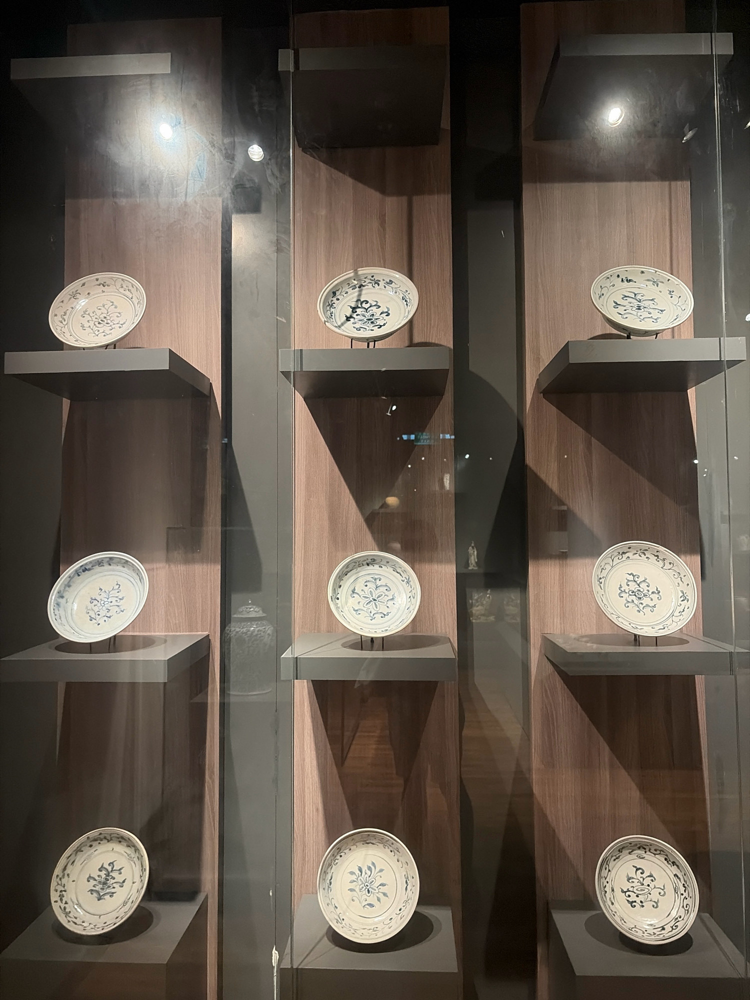 The image shows a display of blue and white porcelain plates in a museum exhibit. The plates are arranged on shelves within glass display cases, with a dark wooden background. The lighting in the exhibit highlights the plates and casts shadows on the wall. It is unclear if this is in Da Nang, Vietnam or if there are any people present.