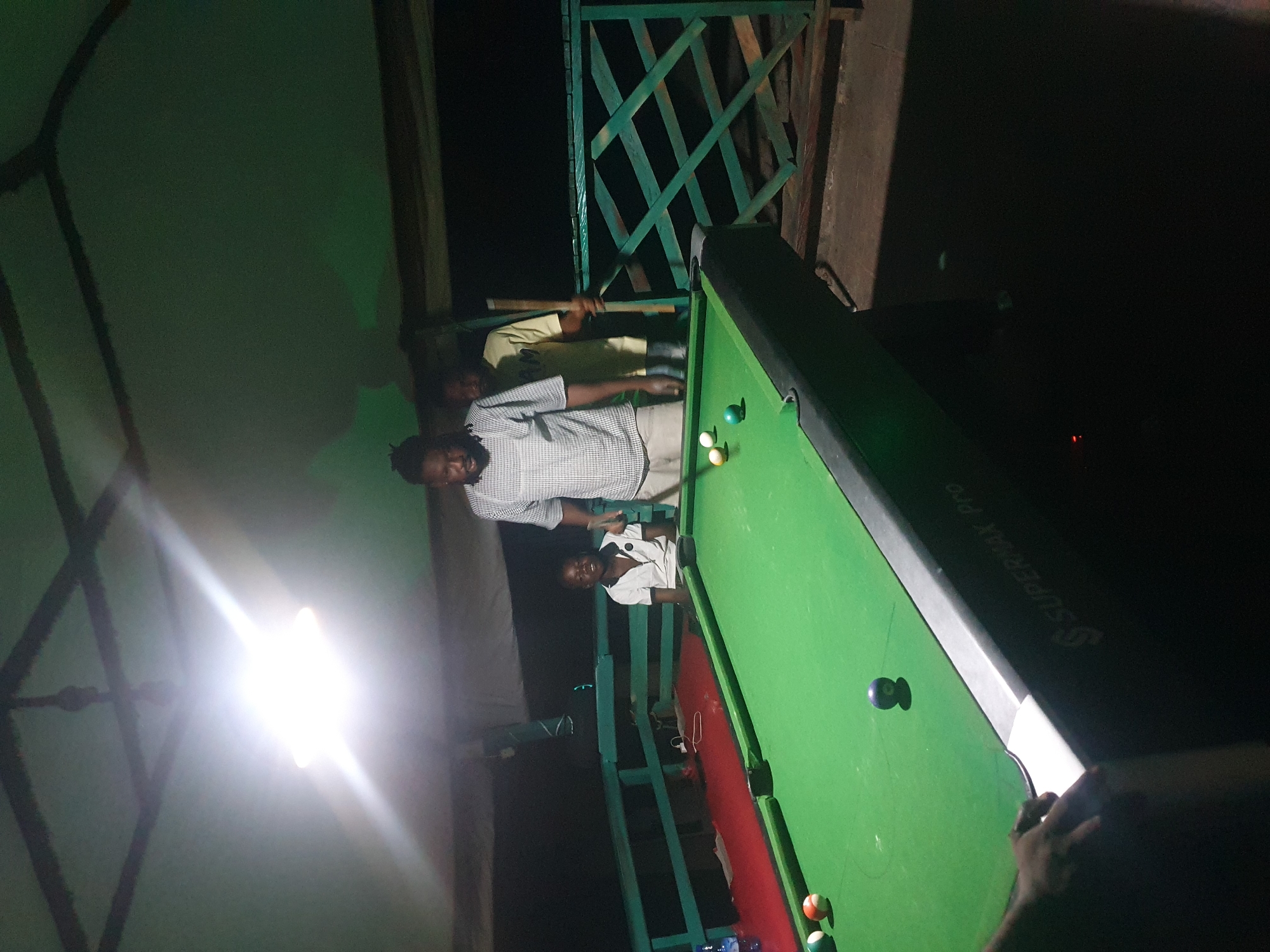 An outdoor covered area, possibly a bar or a recreational space, in Lokoja, Nigeria, features a group of people playing billiards. A bright overhead light illuminates the scene, casting shadows. A man in a checkered shirt stands to the right of a billiard table, holding a cue stick, appearing to play. A young boy in a white polo shirt is seated and smiling at the table. Another man in a yellow t-shirt stands behind the boy, also near the table. The billiard table is green with several balls on it. The surrounding environment includes a wooden railing and a dark, indistinct background.