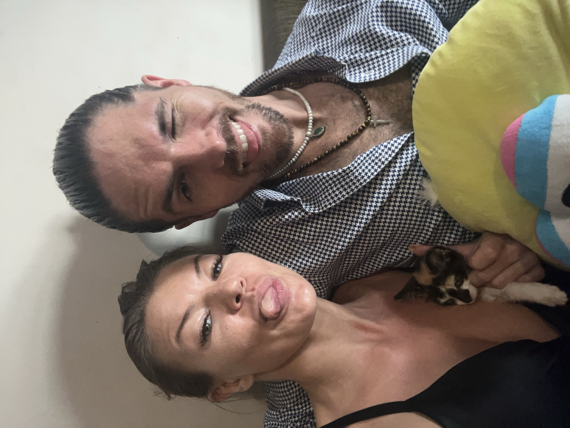 The image depicts a selfie of a man and a woman. The man, located at the top of the frame, winks at the camera and smiles. He has short, dark hair and a beard, and wears a patterned shirt and multiple beaded necklaces.
The woman, positioned below the man, sticks her tongue out. She has dark hair, and wears a black top. Between the woman and the man's chest, lies a small kitten. A yellow pillow is partially visible behind the man. The environment suggests an indoor setting, possibly a domestic interior. There is no information to confirm this picture was taken in Kuta, Indonesia.
