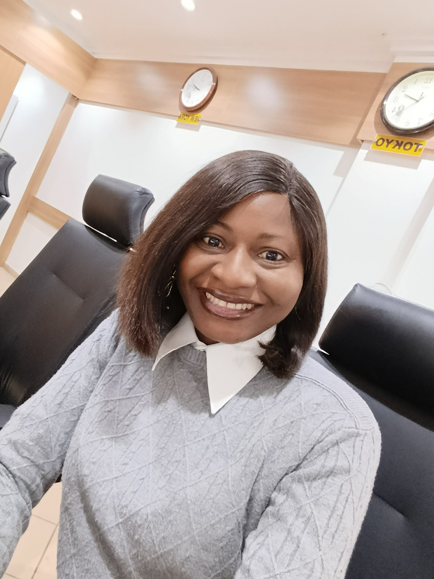 An indoor, close-up photograph depicts a woman smiling directly at the camera while seated in a black or dark grey upholstered chair. She has a medium-brown skin tone and shoulder-length, dark brown hair styled with a side part. She is wearing a light grey long-sleeved sweater with a textured diamond pattern over a white collared shirt. Her expression is a broad smile, revealing her upper teeth.
The background indicates an indoor environment, likely an office or waiting area, within the Immigration Quarters, Nigeria. The walls are white, topped by a light brown wooden paneling accent. Two circular analog wall clocks are mounted on this wooden paneling. Below each clock is a yellow rectangular sign with black, inverted text. The sign below the partially visible left clock reads "NEW YORK" (as "KROY WEN" when viewed directly). The sign below the right clock reads "TOKYO" (as "OYKOT" when viewed directly). Overhead, white recessed ceiling lights provide illumination. Parts of additional black upholstered chairs are visible to the left and right of the woman.