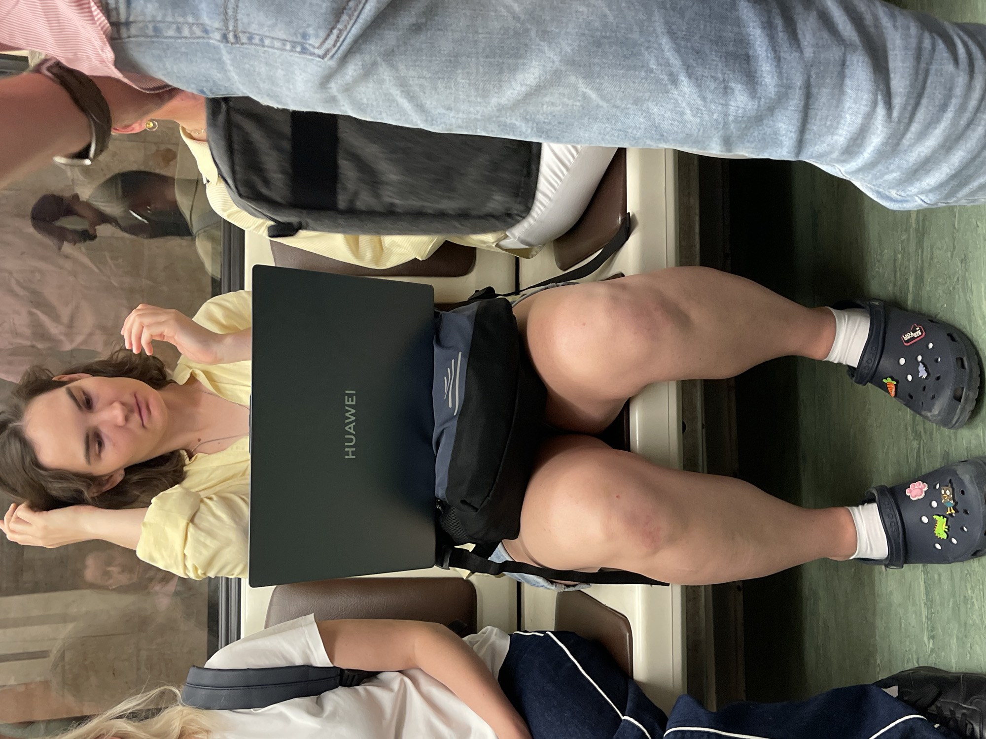 The image captures a scene inside a Moscow Metro car during daylight. The primary subject is a young woman with medium-length brown hair, wearing a light yellow blouse and denim shorts. She is seated, holding an open dark-colored Huawei laptop on her lap. A black fanny pack is worn around her waist, partially obscured by the laptop. She is wearing dark blue Crocs with various colorful Jibbitz charms, including a pink flower and a dinosaur. She appears to be looking down at the laptop, with her right hand touching her hair.
To her left, another young woman with blonde hair is partially visible, wearing a white t-shirt and dark blue track pants with white stripes. She has a dark backpack with black straps on her left shoulder.
To the right of the primary subject, the lower half of a person wearing light blue jeans and a pink and white striped shirt is visible, suggesting they are standing or sitting very close. This person is wearing a dark watch on their left wrist.
The seating in the metro car is upholstered in brown and white material. The background shows the interior of the metro car, with reflections visible in the window behind the subjects, depicting other passengers and the tunnel. The flooring of the car is a green linoleum. The overall lighting suggests it is daytime.