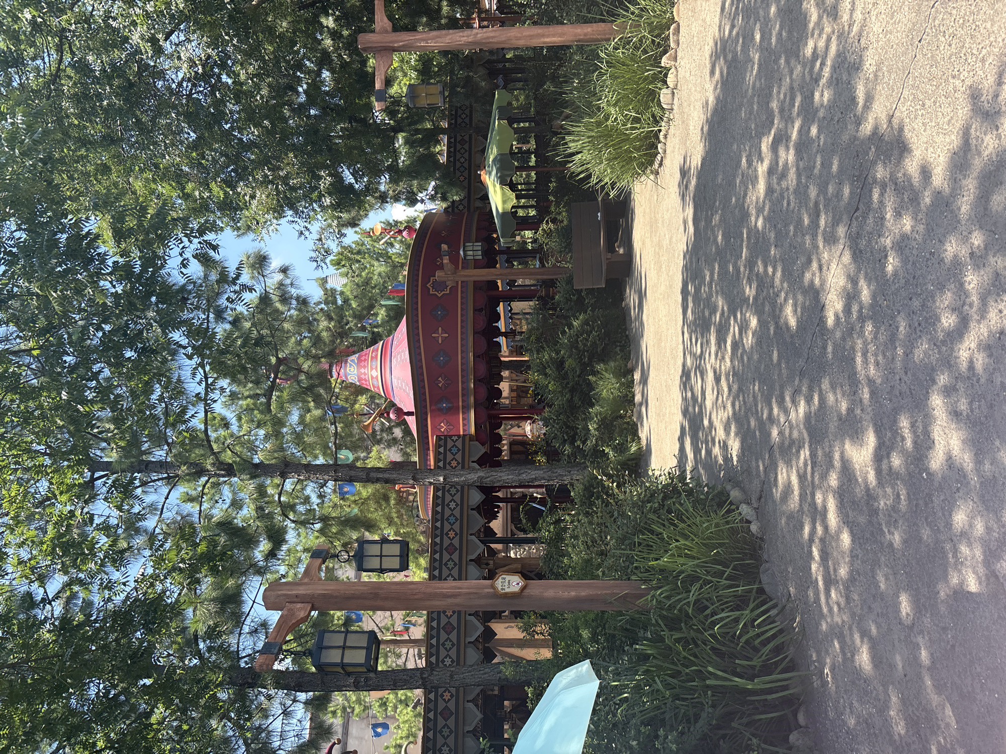 A bright, sunlit outdoor scene features lush green trees with a clear blue sky above. In the mid-ground, a vibrant red and pink carousel structure with intricate decorative patterns stands prominently. Several wooden posts with hanging lanterns are strategically placed throughout the scene. A light blue umbrella is partially visible on the left, and a brown wooden bench is situated near a green and yellow umbrella on the right. The ground is covered with a light-colored gravel path, dappled with shadows from the surrounding foliage. The image captures a lively and whimsical atmosphere, likely in a park or amusement area. There is no identifiable signage or landmarks to confirm a specific location in Pudong, China.