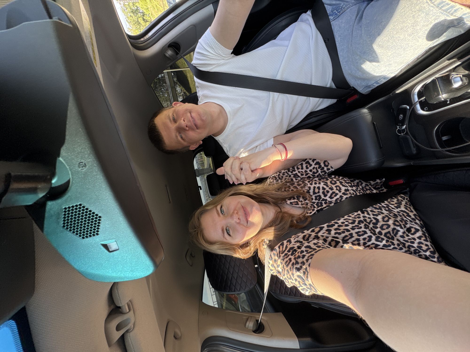 Inside a car, a fair-skinned woman with blonde hair, wearing a leopard-print top and a seatbelt, sits next to a fair-skinned man with short brown hair, wearing a white shirt, jeans, and a seatbelt. They are holding hands. The interior of the car includes black leather seats, a rearview mirror, a sunroof, and the car's console area with keys and a gear shift. The photo appears to be taken from a slightly elevated perspective. Based on the license plate, the car may be registered in the city of Perm, Russia.