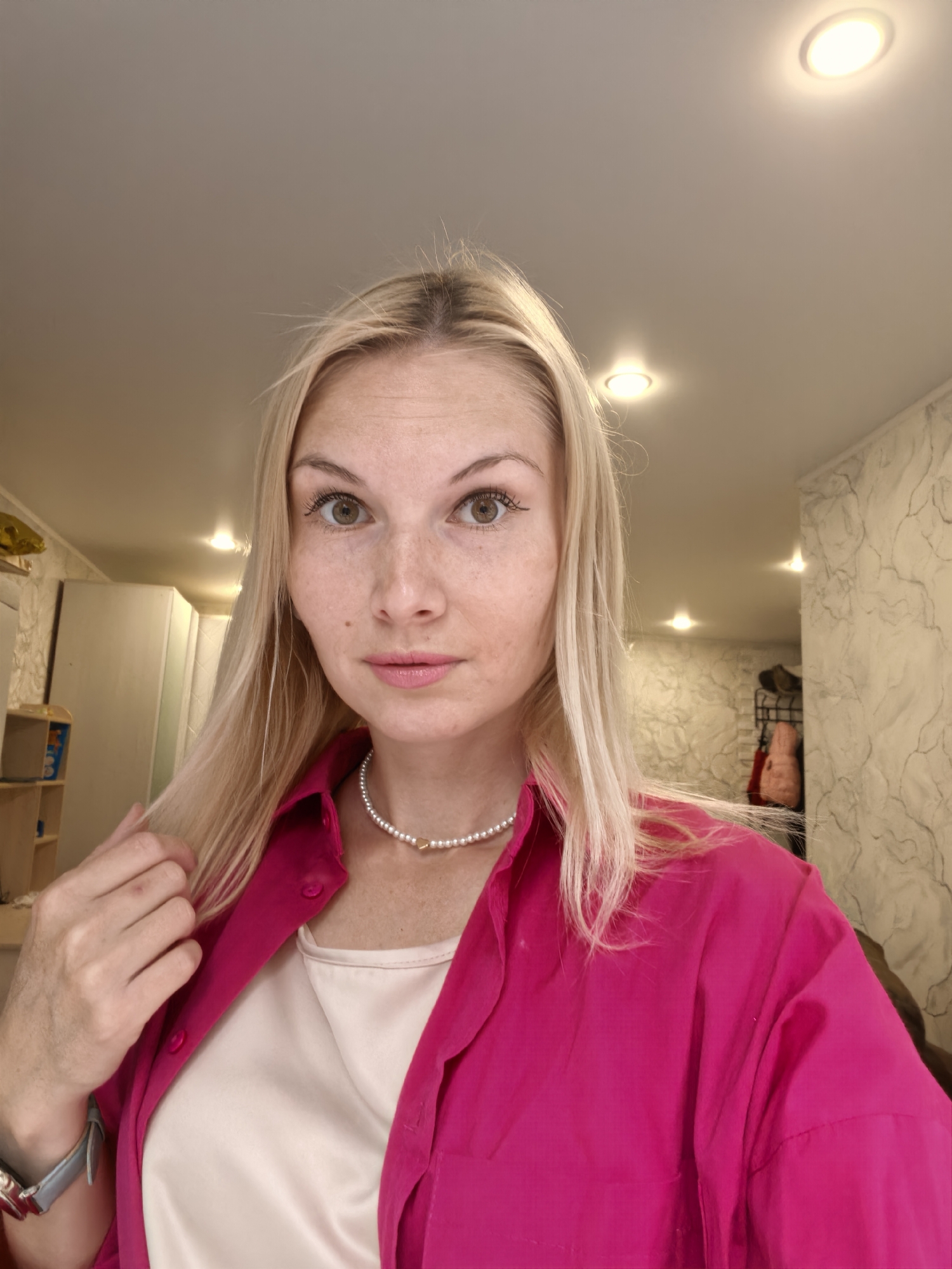 A young woman with blonde hair and light skin is taking a selfie in what appears to be an indoor setting, possibly a room in Sterlitamak, Russia. She is wearing a bright pink button-up shirt over a light-colored camisole, and a pearl necklace with a small gold heart pendant. Her left hand is near her hair, and she has a watch on her left wrist. The background shows a ceiling with recessed lighting, a wall with a textured wallpaper pattern, and some furniture, including cabinets and a shelf unit with toys. The lighting in the room is bright, likely from the overhead lights.