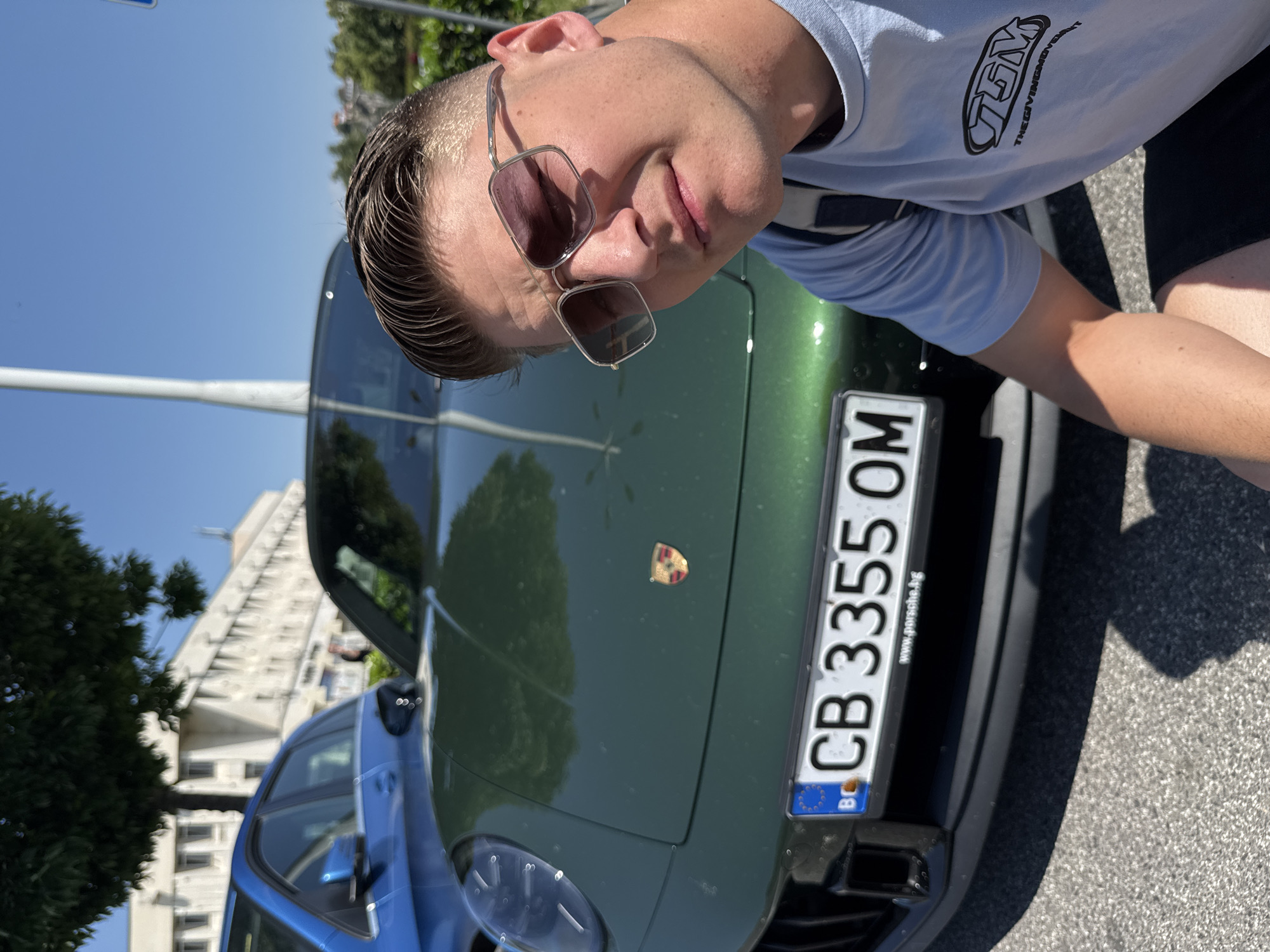 A man in sunglasses and a light blue t-shirt with the logo "TGM THEGIVINGMOVEMENT" is posing for a selfie in front of a green Porsche. The car has a Bulgarian license plate reading "CB 3355 OM". Another blue car is partially visible in the background. A building is visible in the background. The location is the city of Plovdiv, Bulgaria.