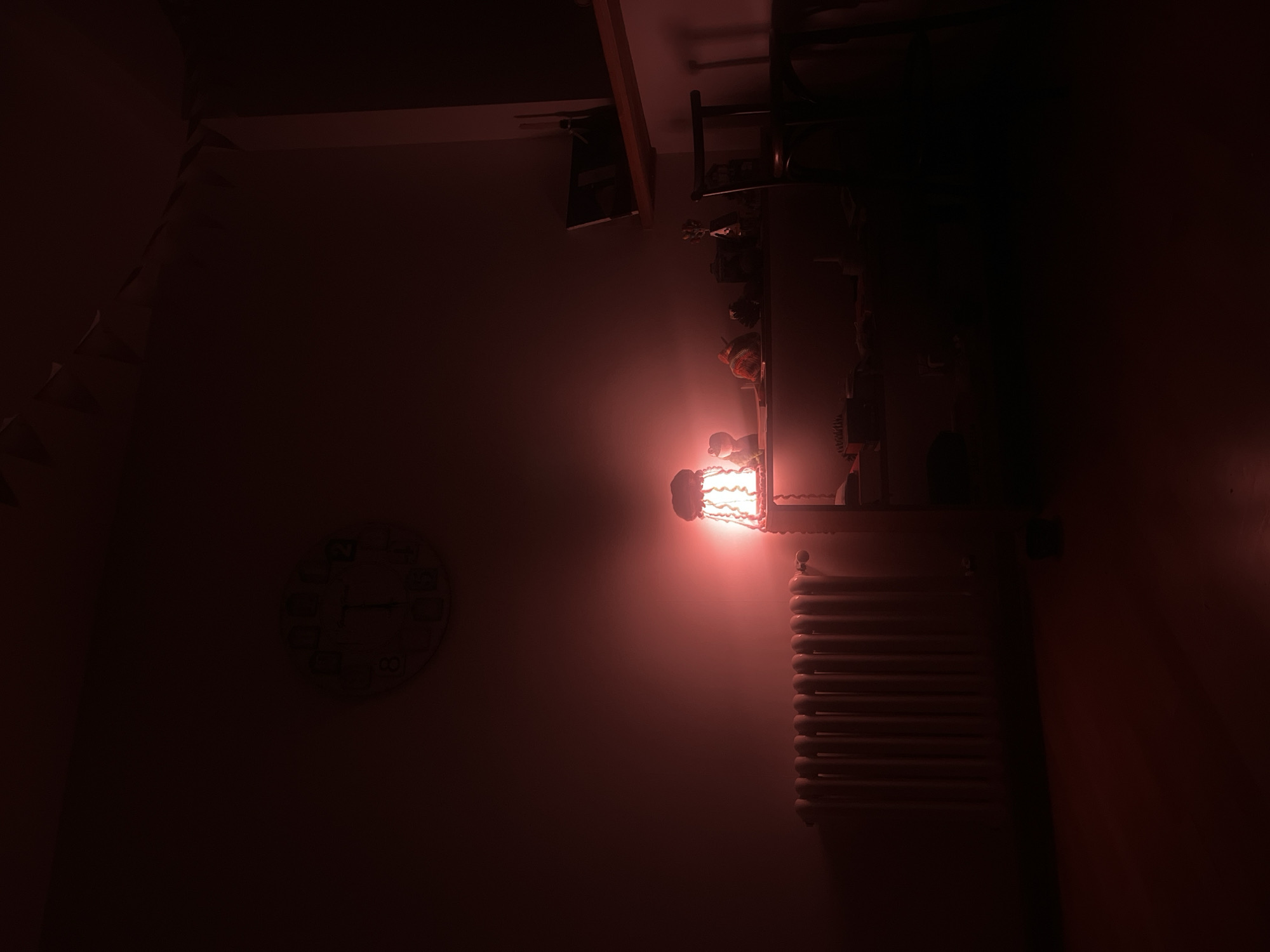 The image depicts a dimly lit room. A radiator is visible on the right side of the frame, illuminated by a bedside lamp above it. To the left of the radiator, on the wall, is a clock. The location appears to be inside an unknown room, in an unknown city. The scene seems to be indoors and at night.