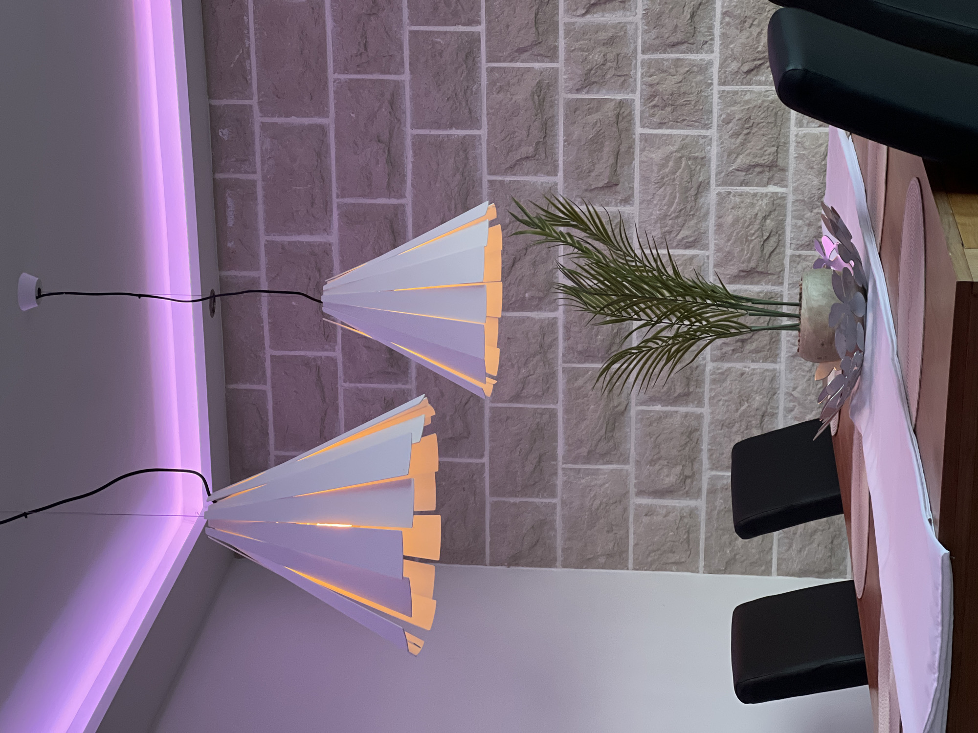 The image shows an interior scene in Ivanovići, Montenegro. Two white, conical pendant lamps with warm-toned lighting elements hang from the ceiling. One lamp has a visible power cable leading to a white wall-mounted electrical outlet. The ceiling features a strip of purple LED lighting along the edge. A brick-patterned wall serves as the backdrop. A green plant in a pot is located near the brick wall. Part of a dining table, a light pink table runner, and two black chairs are visible in the foreground. A black leather chair is also visible in the upper right corner.