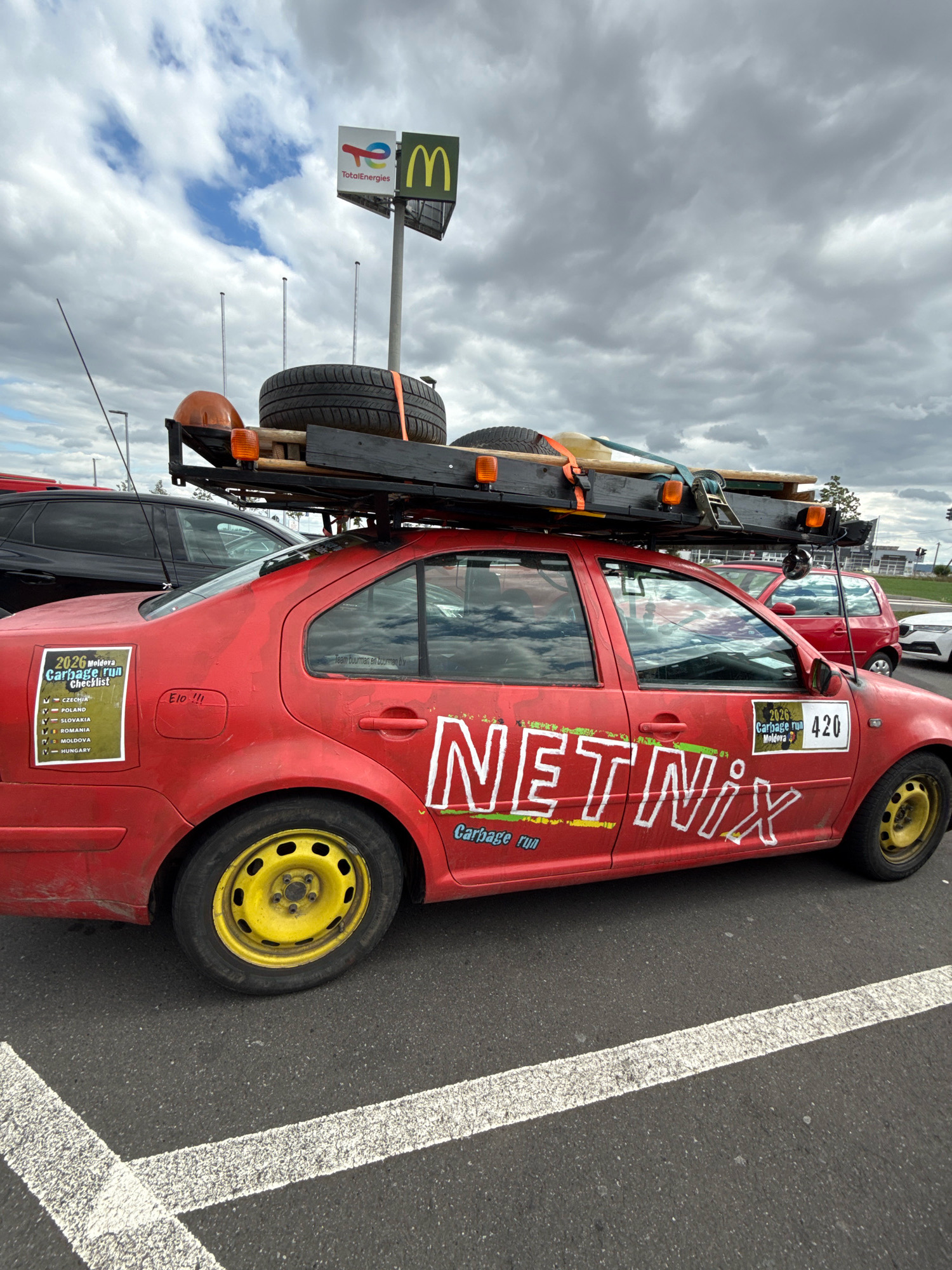 A red sedan, a Volkswagen Passat, with "NETNIX Garbage Run" written in white, stylized lettering across its side, is parked in a lot with white painted lines. The car has yellow rims and a spare tire and what appears to be a ladder strapped to its roof rack. A sign with the logos for TotalEnergies and McDonald's is visible in the background. The scene is overcast with clouds. The car is parked in a lot at a McDonald's in Werther, Germany.