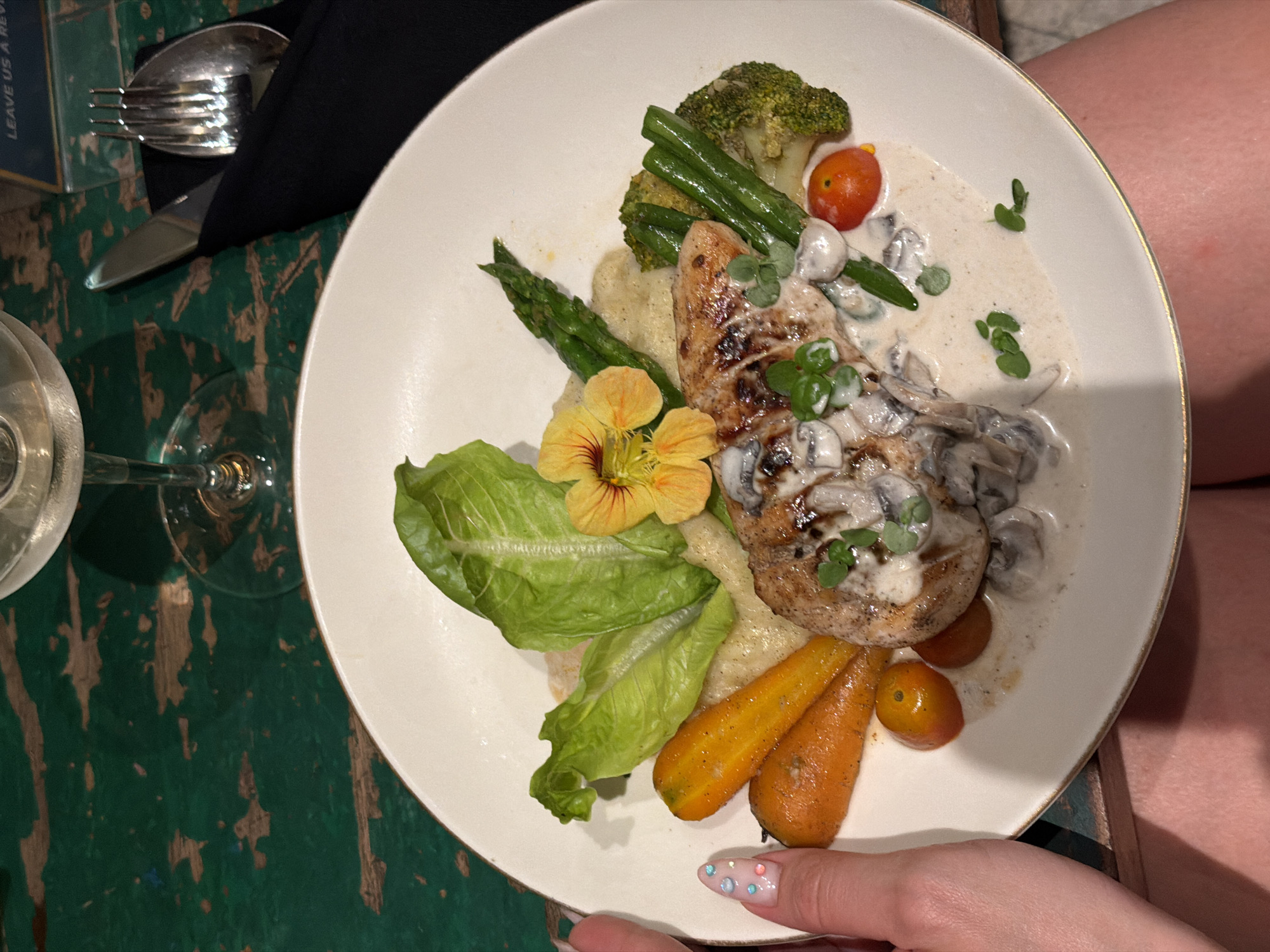 A top-down shot shows a prepared meal on a white plate with a gold rim, set on a distressed green wooden table. The meal consists of grilled chicken topped with a creamy mushroom sauce, accompanied by roasted carrots, green beans, asparagus, broccoli florets, and a single cherry tomato. A nasturtium flower and lettuce leaves are used as garnish. A glass of white wine sits to the left of the plate. To the upper left of the frame, a black napkin is folded beside a fork and knife. The background is blurred, but a person's legs are visible to the right, suggesting they are seated at the table. The lighting appears to be natural, indicating it might be daytime. The overall mood is relaxed and inviting, suitable for a dining experience. There is a small white label visible on the upper left with text that is partially cut off, but the words "LEAVE US A REVI" can be discerned.