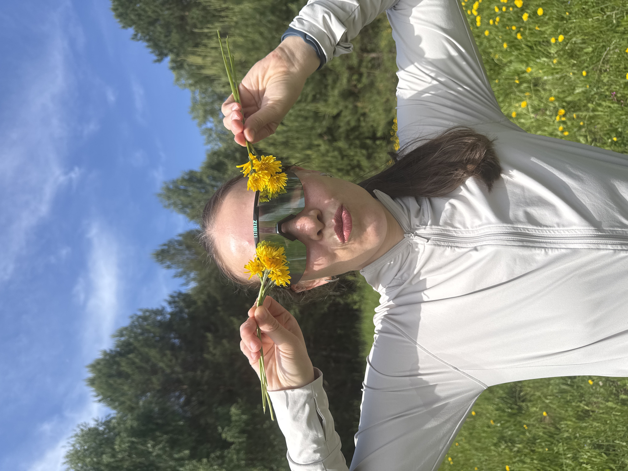 The image displays a person, likely a woman, outdoors in a bright, natural setting in Chaikovsky, Russia. The person is wearing a light gray, long-sleeved athletic top with a zippered collar and large, reflective sports sunglasses. The word "BLIZ" is visible on the side frame of the sunglasses.
The central activity shows the person holding small bouquets of bright yellow dandelions, positioning them playfully near their sunglasses, almost as if creating a floral frame or decoration around their eyes. The background is a vibrant green field or meadow, densely covered with blooming yellow dandelions. Further in the background, there are lush green trees, indicating a forested or natural area. The sky above is a clear, bright blue with a few wispy white clouds, suggesting a sunny day, likely late morning or afternoon. The overall impression is one of natural beauty and lightheartedness.
