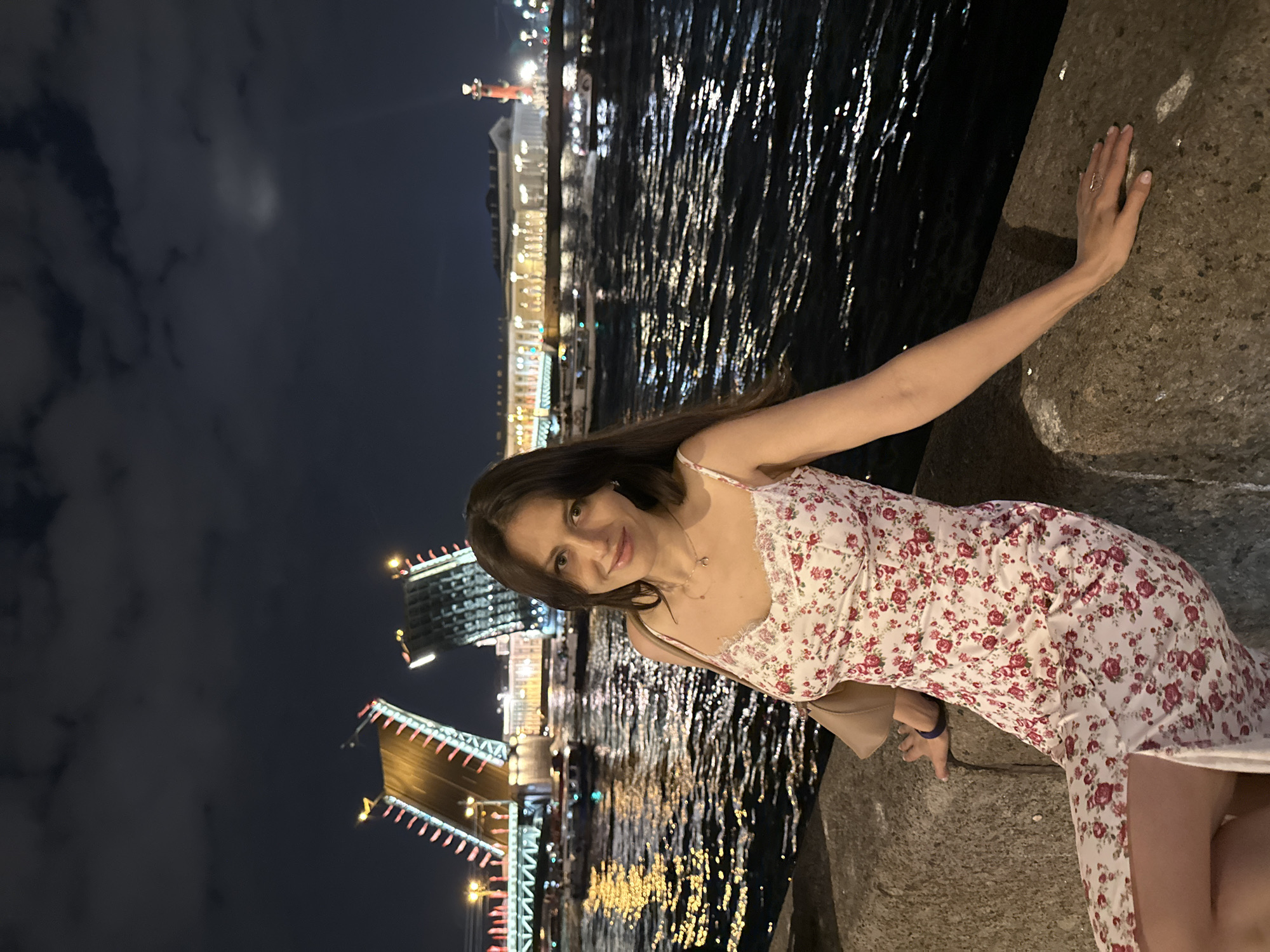 A medium shot captures a woman posing against a low wall or embankment at night. She is wearing a white, floral dress with red and pink patterns. Her hair is long and dark, and she is wearing a necklace. She is leaning against the wall with one hand, her other hand visible at the waist.
In the background, there is a large body of water reflecting city lights. The Peter and Paul Fortress is partially visible, its spire illuminated. A bridge, possibly the Palace Bridge, is raised in the open position, its lights bright. The scene suggests a tourist location in the city of Saint Petersburg, Russia, at night.