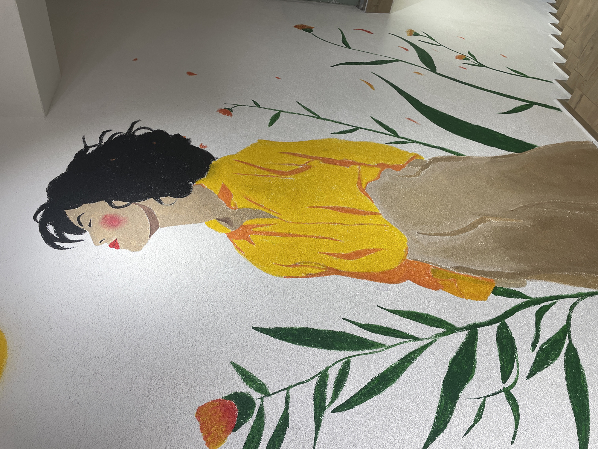 The image is a photograph of a mural painted on a white wall, likely in Kadıköy, Turkey. The mural depicts a stylized young woman with dark, curly hair, her head tilted back with eyes closed and a serene expression. She is wearing a bright yellow collared shirt and a tan skirt or trousers. To the left of the woman, a large orange and red flower with green leaves is painted, and to the right, several thin green stems with small orange flowers and scattered red and orange petals are visible. A faint yellow circle, possibly representing the sun, is partially visible in the upper left corner of the mural. The wall has a textured surface. The bottom right of the image shows a portion of wooden stairs and a white baseboard with a jagged edge.