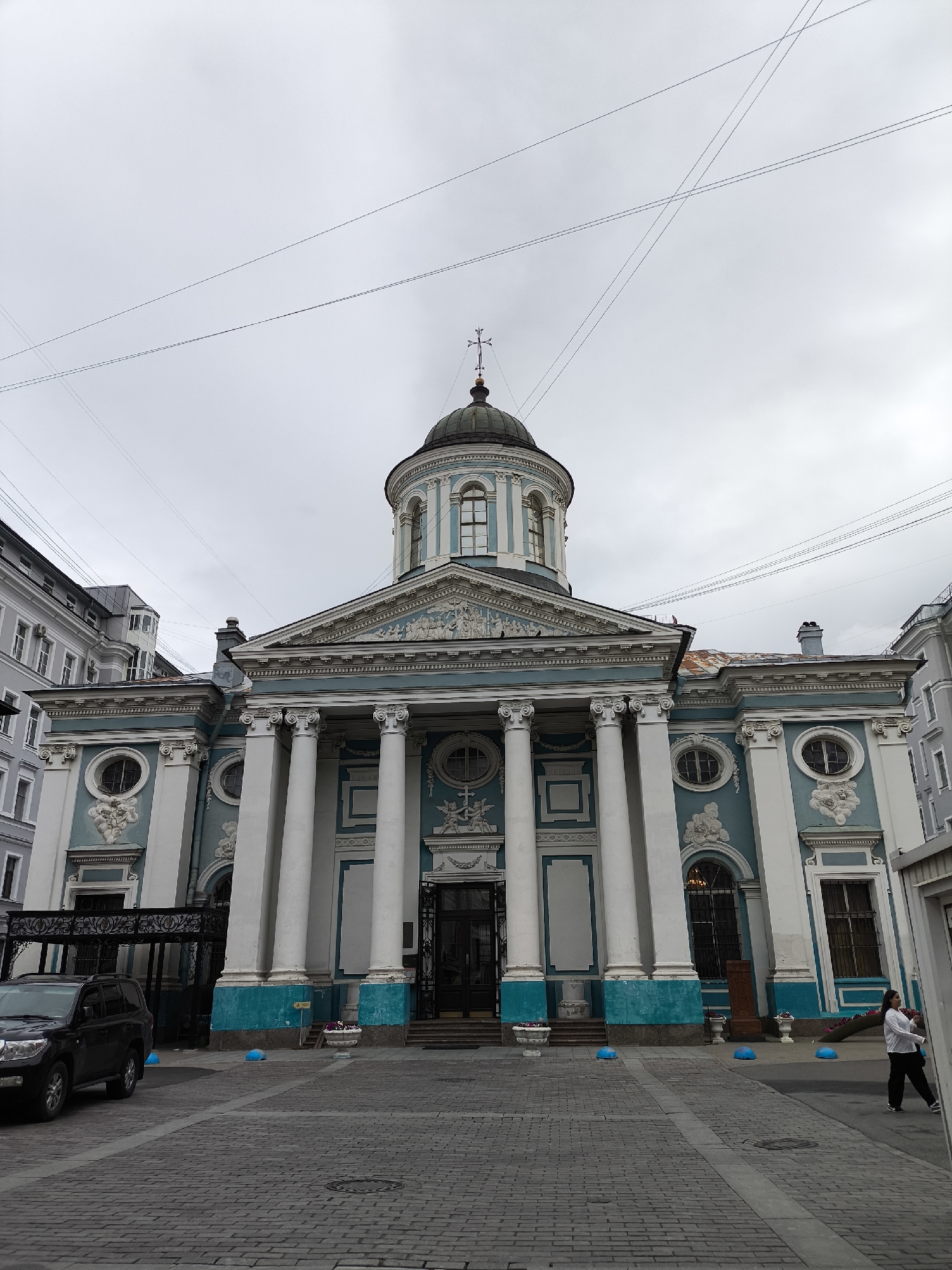 The image shows the Church of the Assumption of the Blessed Virgin Mary, also known as the Catholic Church of the Assumption, located at 35-37 Rimsky-Korsakov Avenue in Saint Petersburg, Russia. The church is a blue and white Neoclassical building with a dome and columns. It has a large portico with white columns supporting a pediment. The building is adorned with decorative elements and has large windows. In front of the church, there is a paved courtyard with a black SUV parked on the left. A few blue bollards are visible in the foreground. A woman in a white shirt and dark pants is walking on the right side of the frame. The sky is overcast and cloudy. The surrounding buildings are also visible, with one to the left appearing to be an apartment building with a gray facade.