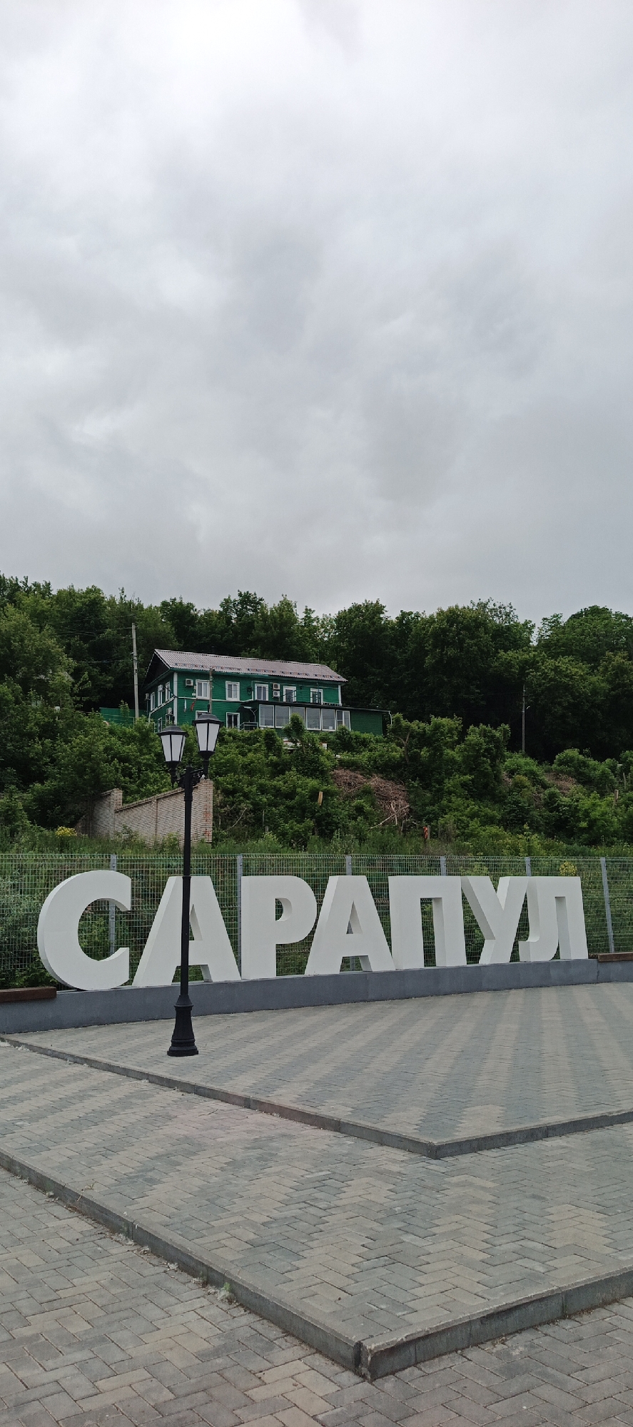 The image presents a static, outdoor scene in Sarapul, Russia, on an overcast day. A large, white, three-dimensional sign spelling "САРАПУЛ" in Cyrillic dominates the lower-midground, resting on a dark grey base. Behind the sign, a light green mesh fence separates it from a densely forested hillside. A classic-style black lamppost with two unlit luminaires stands in the lower-center, partially obscuring the initial letters of the sign.
On the hillside, a two-story building with dark green siding, a brown roof, and multiple white-framed windows is visible. The building features a ground-floor extension with large glass windows. The surrounding hill is covered with lush, dark green deciduous trees and shrubs, with a light-colored brick wall section discernible on the left side of the hill. A utility pole with wires is partially obscured by trees on the right side of the hill.
The sky in the upper portion of the image is light grey and heavily overcast, with visible cloud formations. The foreground consists of an expanse of paving stones laid in a herringbone pattern, using grey and light brown rectangular pavers. Raised concrete curbs delineate distinct sections of the paved area, forming a pathway or plaza. No people are present in the scene.