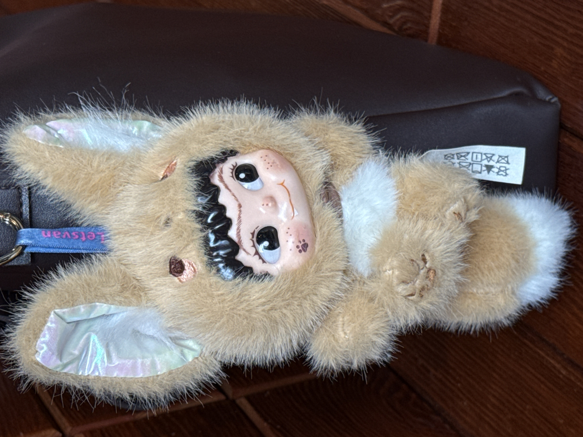 A fluffy plush toy, possibly a keychain, is depicted in a close-up shot, lying on its back on a dark brown, slightly textured surface, which appears to be a bag or cushion. A wooden surface with visible grain is present in the background and extends into the foreground. The image itself is rotated approximately 90 degrees clockwise.
The toy is primarily covered in shaggy, light brown or tan fur, with patches of white fur on its body, particularly on its chest or belly area. Its face is made of a smooth, light-colored material, likely plastic or ceramic, and is embedded within the fur. The face features large, dark, glossy eyes with white highlights, small dark red lips forming a slight pout, and several faint freckles on the nose and forehead. A small, dark paw print design is visible on its right cheek. Black, curly, molded hair frames its face. Two prominent ears extend from its head; the inner surfaces of these ears are made of a shiny, iridescent material that reflects hues of pink, green, and blue. A small, embroidered brown heart or similar shape is located on the left side of the toy's face.
Attached to the toy is a blue fabric ribbon loop with the word "Letsvan" printed on it in white capital letters. A small white label, partially obscured by fur, is visible near the toy's body. This label displays common laundry care symbols, including "do not wash," "do not bleach," "do not tumble dry," and "do not iron," among others. There is no discernible activity taking place; the image is a still photograph of the object. The lighting suggests an indoor setting, likely during the day.