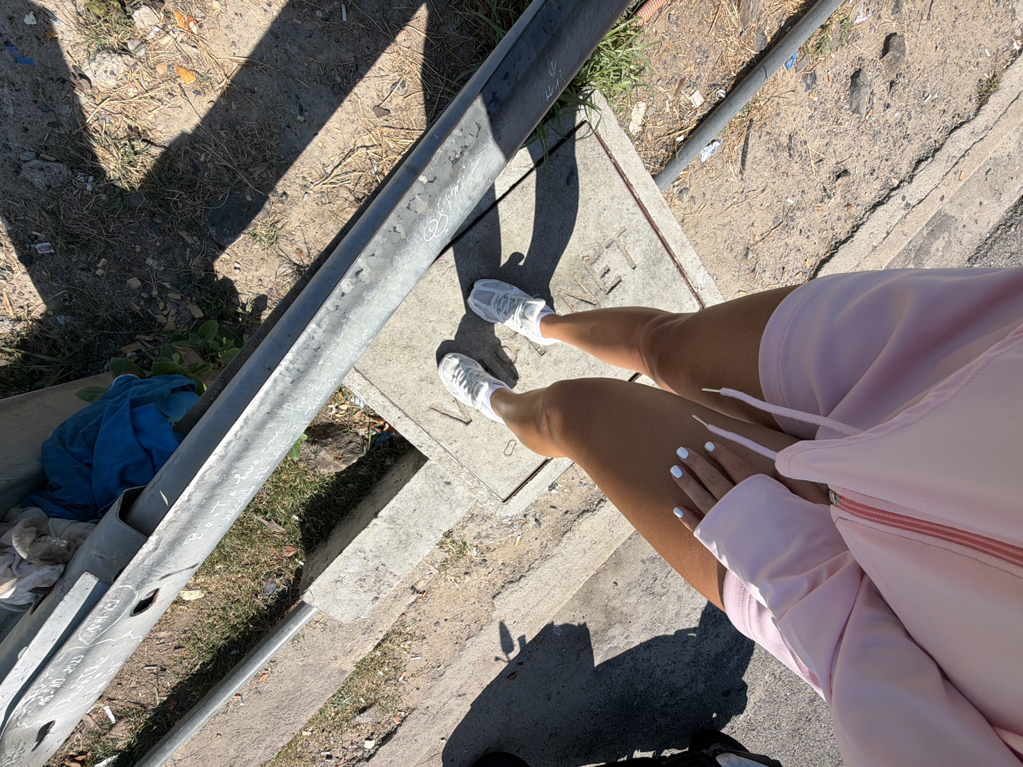 A person wearing white sneakers and a pink tracksuit is standing outdoors. The person's legs and arms are visible, along with their hands, which have light blue nail polish. The ground beneath them consists of concrete slabs and dry grass, with shadows cast by sunlight. A metal pipe runs horizontally across the scene, and some blue and white fabric is draped over it. The scene appears to be in an urban outdoor environment. There is no specific indication of the location being Nam Nha Trang Ward, Vietnam.