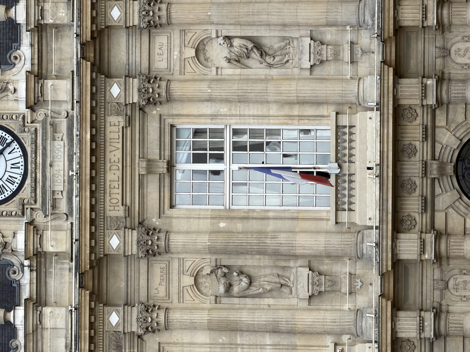 This is a frontal, medium shot of the Hôtel de Ville (City Hall) in Ivry-sur-Seine, France, a civic building constructed in stone. The architecture features classical elements such as columns, ornate carvings, and a large, multi-paned window with a French flag hanging from it. Above the window, the text "HOTEL-DE-VILLE" is carved into the facade. To the left of the window, a niche contains a statue of a man holding grapes, with the inscription "LA PIERRE" above it. To the right, another niche holds a statue of a man with a tool, and the inscription "L'EAU." The building has a clock face visible at the very top of the frame, and the years "1894-1896" are inscribed below it, likely indicating the construction dates. The overall impression is one of grandeur and historical significance.
