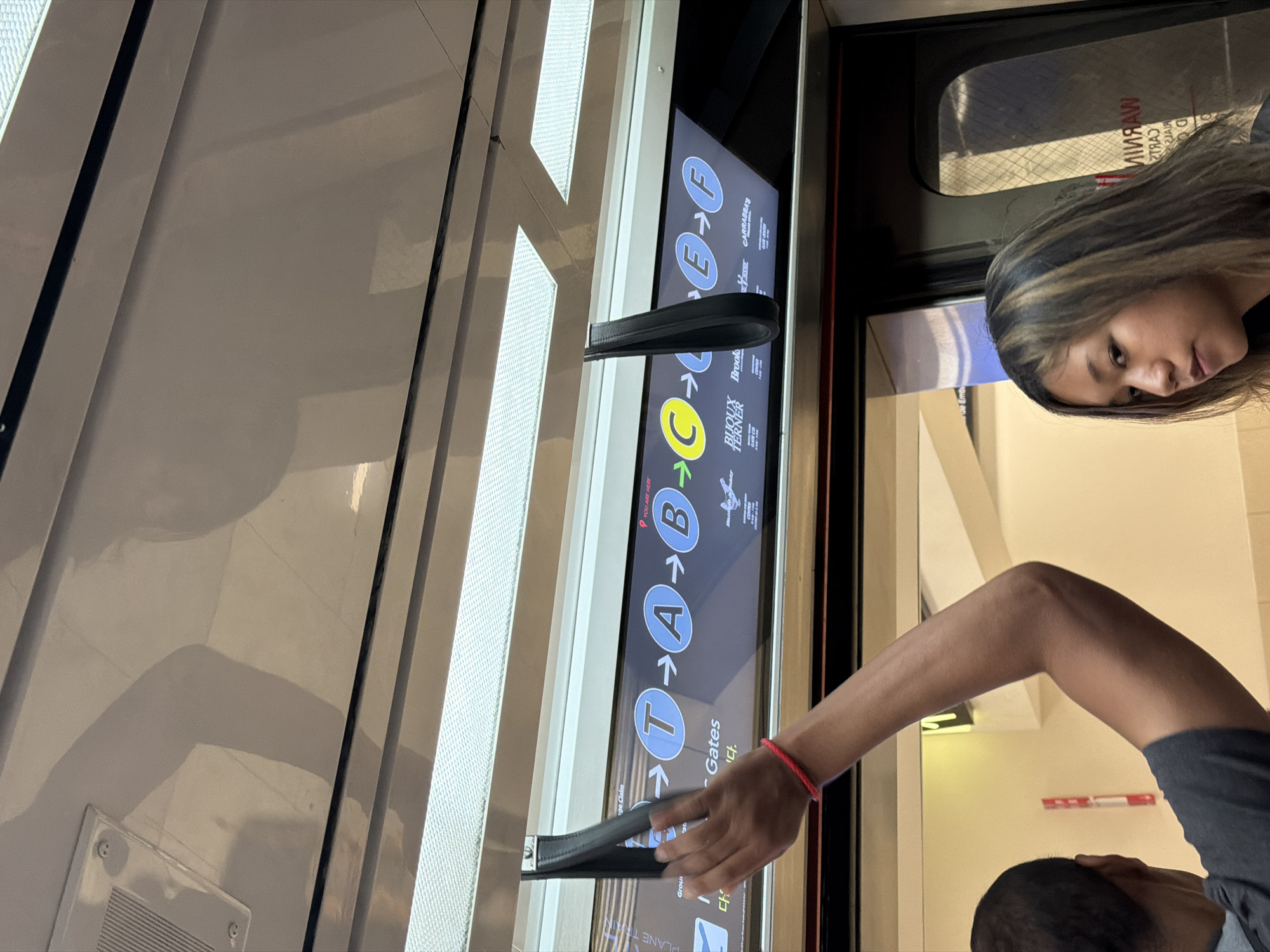 This image shows an interior view of a train, likely part of the Plane Train system at Hartsfield-Jackson Atlanta International Airport. The train car has a digital display showing a route with letters T, A, B, C, E, and F, indicating different stops or concourses. A person's arm, adorned with a red bracelet, is visible reaching for a black strap handle overhead. In the foreground, a young woman with blonde-streaked hair is looking to her left. The environment appears to be well-lit with fluorescent lighting.