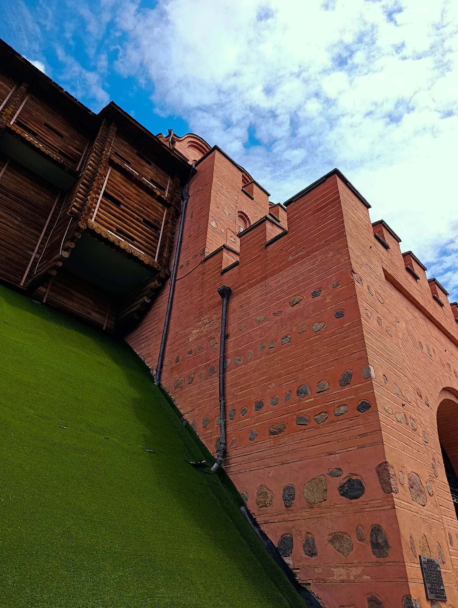 The image showcases a section of the ancient fortress walls in Kyiv, Ukraine. The walls are constructed from red brick and feature stone inlays, with crenellated battlements visible at the top. To the left, a section of a wooden structure, possibly a watchtower or part of the fortress's defensive architecture, is visible. A steep, vibrant green incline, likely artificial turf, slopes downwards from the base of the wall, suggesting a modern park or recreational area built around the historical site. The sky above is a bright blue with scattered white clouds. The perspective is low-angle, emphasizing the height and imposing nature of the fortress.