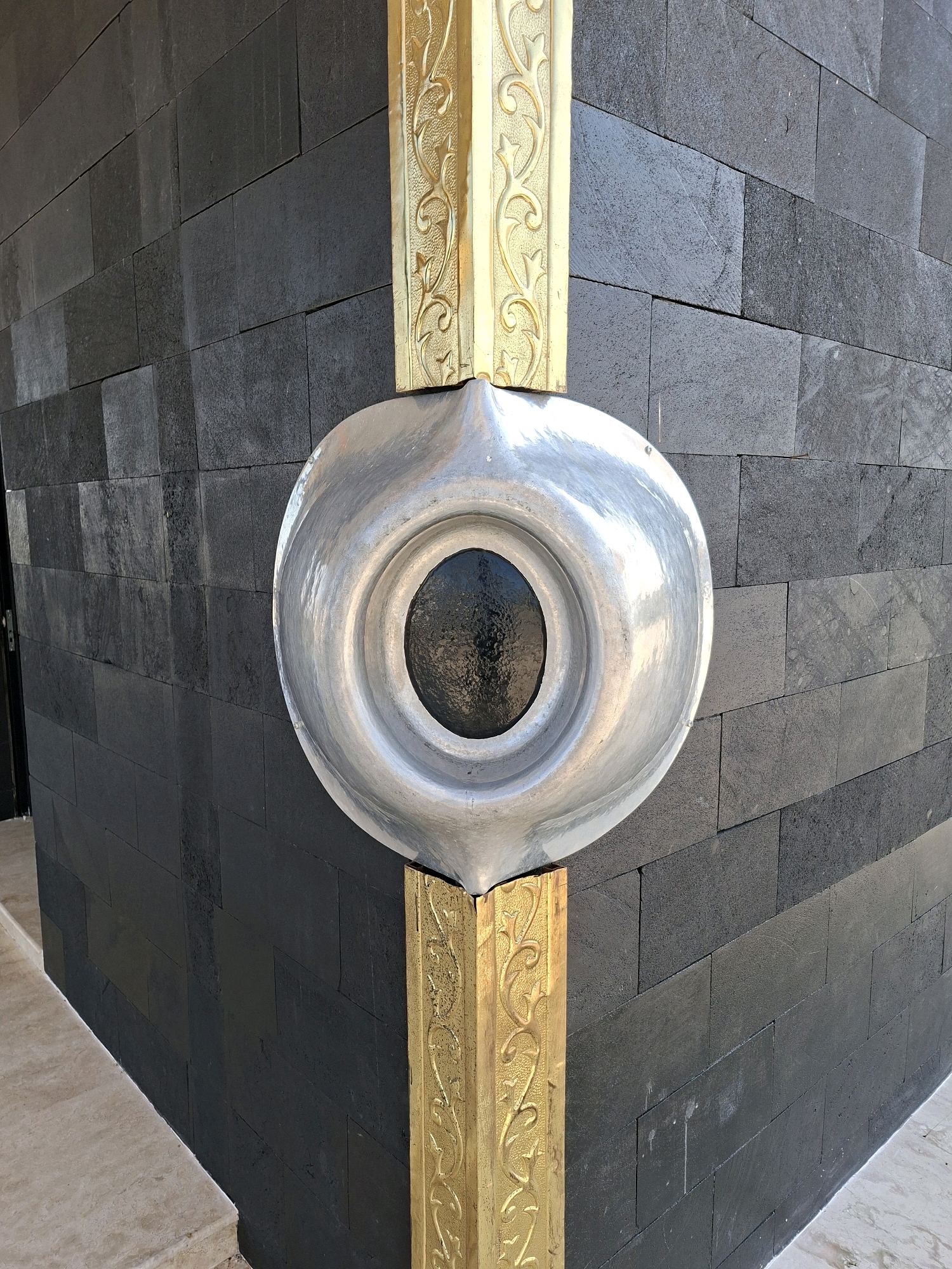 The image shows a close-up of the corner of a building with black stone walls and gold decorative trim. A metallic, oval-shaped object with a dark, circular center is mounted on the corner, partially obscuring the gold trim. The object appears to be made of polished metal and has a textured surface around the central dark area. The building's corner has a sharp angle, and the surrounding area suggests an outdoor environment with paved flooring visible at the bottom. The lighting indicates daylight. The description of the location in Jrengik, Indonesia, is external information not derivable from the visual content of the image itself.