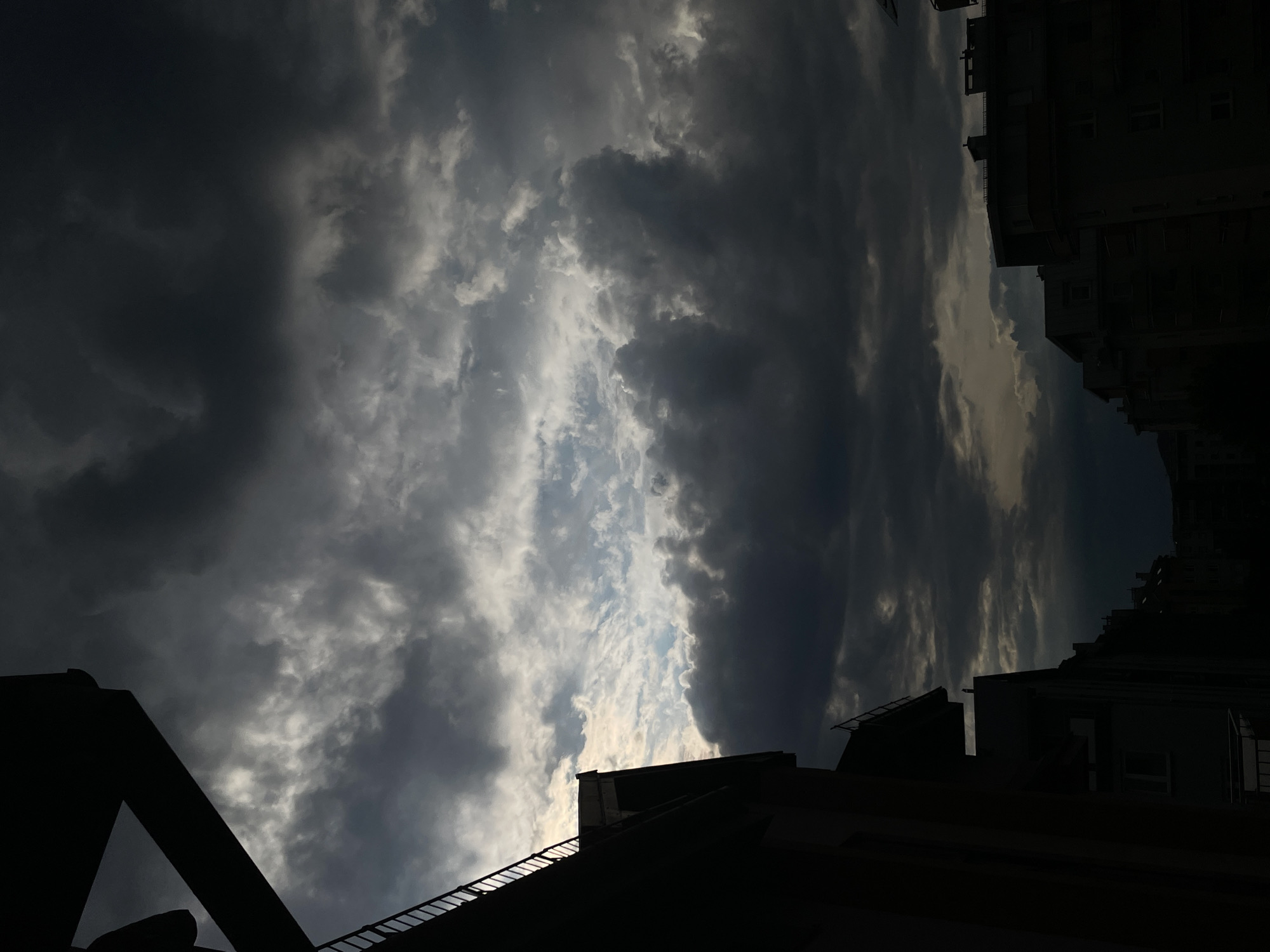 The image captures a city skyline under a dramatic, overcast sky. Dark, heavy clouds dominate the upper portion of the frame, with patches of lighter sky peeking through in the center, suggesting either a break in the clouds or the fading light of day. The silhouettes of buildings are visible along the bottom and right side of the image, with architectural details such as balconies and rooftops discernible despite the low light. The left side of the image features a dark, angular structure, possibly a building or a piece of infrastructure, casting a shadow over the scene and framing the sky. The overall mood is somber and atmospheric, hinting at approaching weather changes or the end of a day. There are no people or discernible actions taking place.