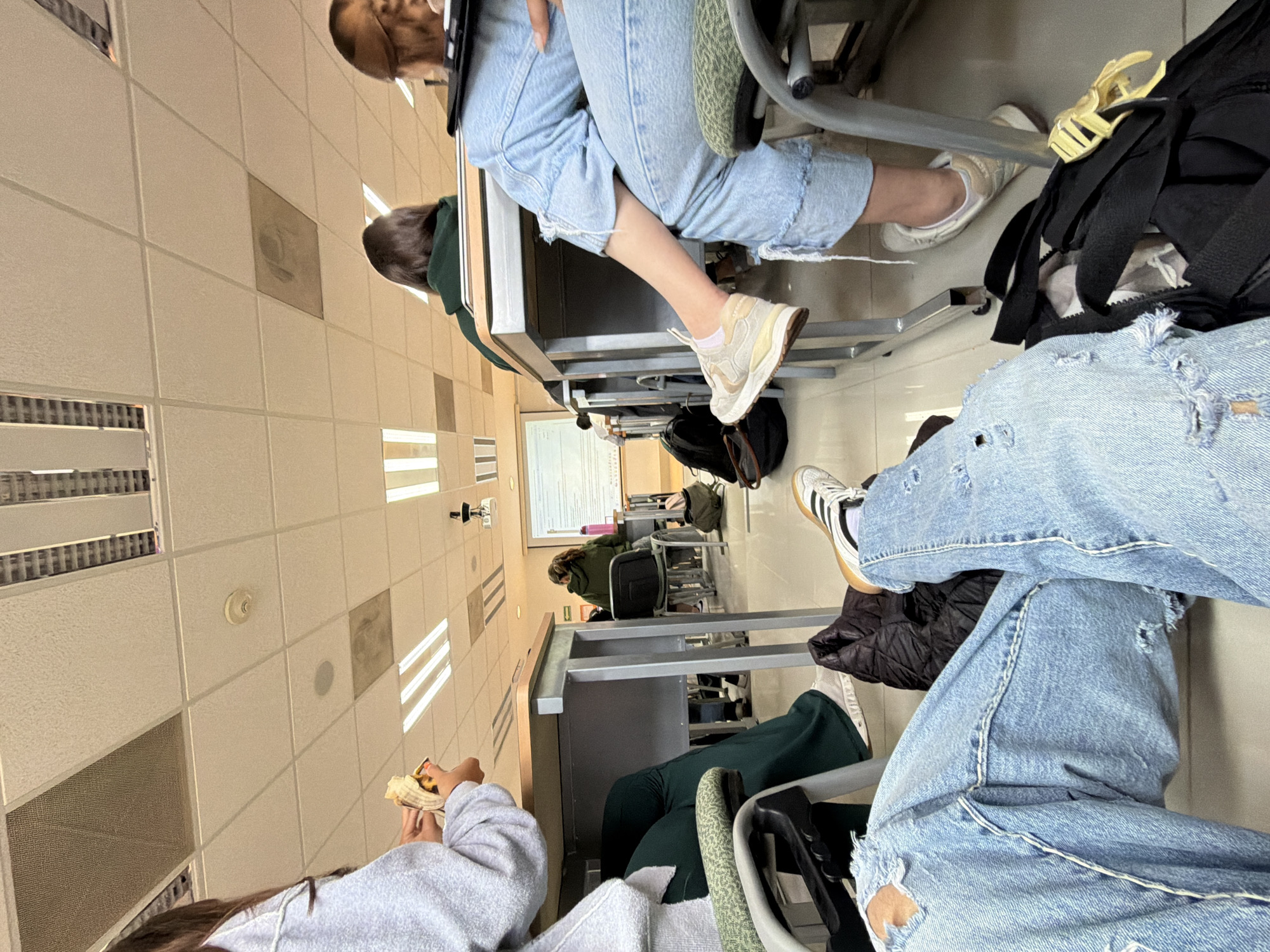 This image captures a low-angle perspective inside what appears to be a classroom or lecture hall, viewed from a seated position. The setting is bright with artificial lighting from overhead fluorescent panels integrated into a grid of ceiling tiles.
In the immediate foreground on the right, a person is seated, wearing light blue ripped jeans with prominent white stitching and white Adidas sneakers with black stripes and a light brown sole. A black backpack with a yellow clip or handle is visible beside them, under the desk.
To the foreground left, another person is seen from the shoulder up, wearing a light grey hoodie and holding what looks like a partially peeled banana or a pastry.
Further into the mid-ground, another individual is seated at a desk, wearing light blue jeans and light beige sneakers with textured soles. Behind them, another person is visible, wearing a dark green top or hoodie. Numerous other desks and chairs extend into the background, with more students partially visible.
At the far end of the room, a white board or projection screen is visible, though its content is indistinct. The room features light-colored walls and a speckled floor. The overall atmosphere is casual, suggesting a break or an informal class setting. There is no discernible text visible in the image. The provided context indicates the location is San Andrés Cholula, Mexico.