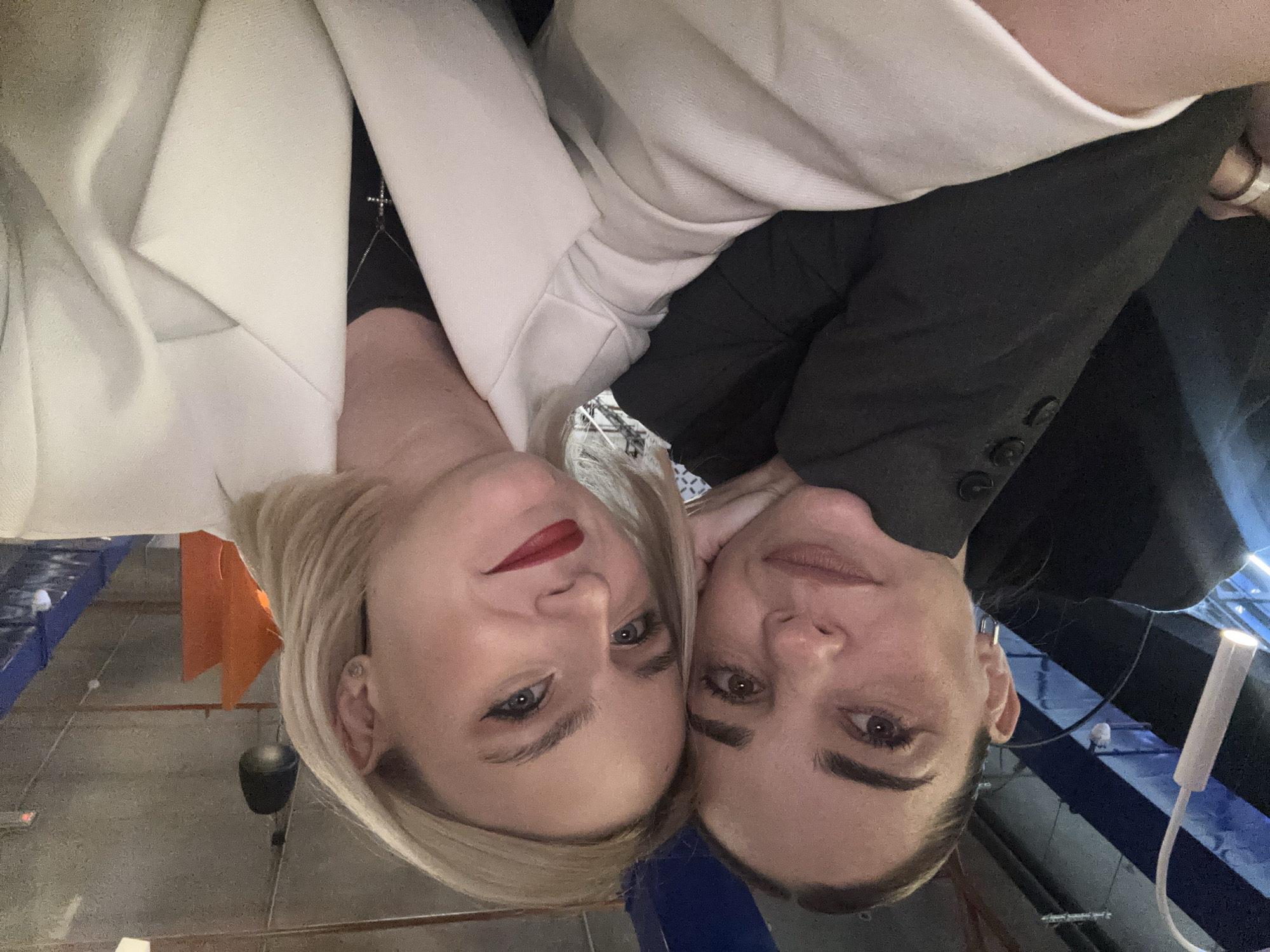Two women are posed in a close-up selfie, heads tilted towards each other. The woman on the left wears a white blazer with a cross necklace visible beneath, and red lipstick. The woman on the right wears a black blazer. The background is a modern, industrial space with blue and gray elements. Overhead lighting fixtures are visible in the background. The location is Unknown, Russia.